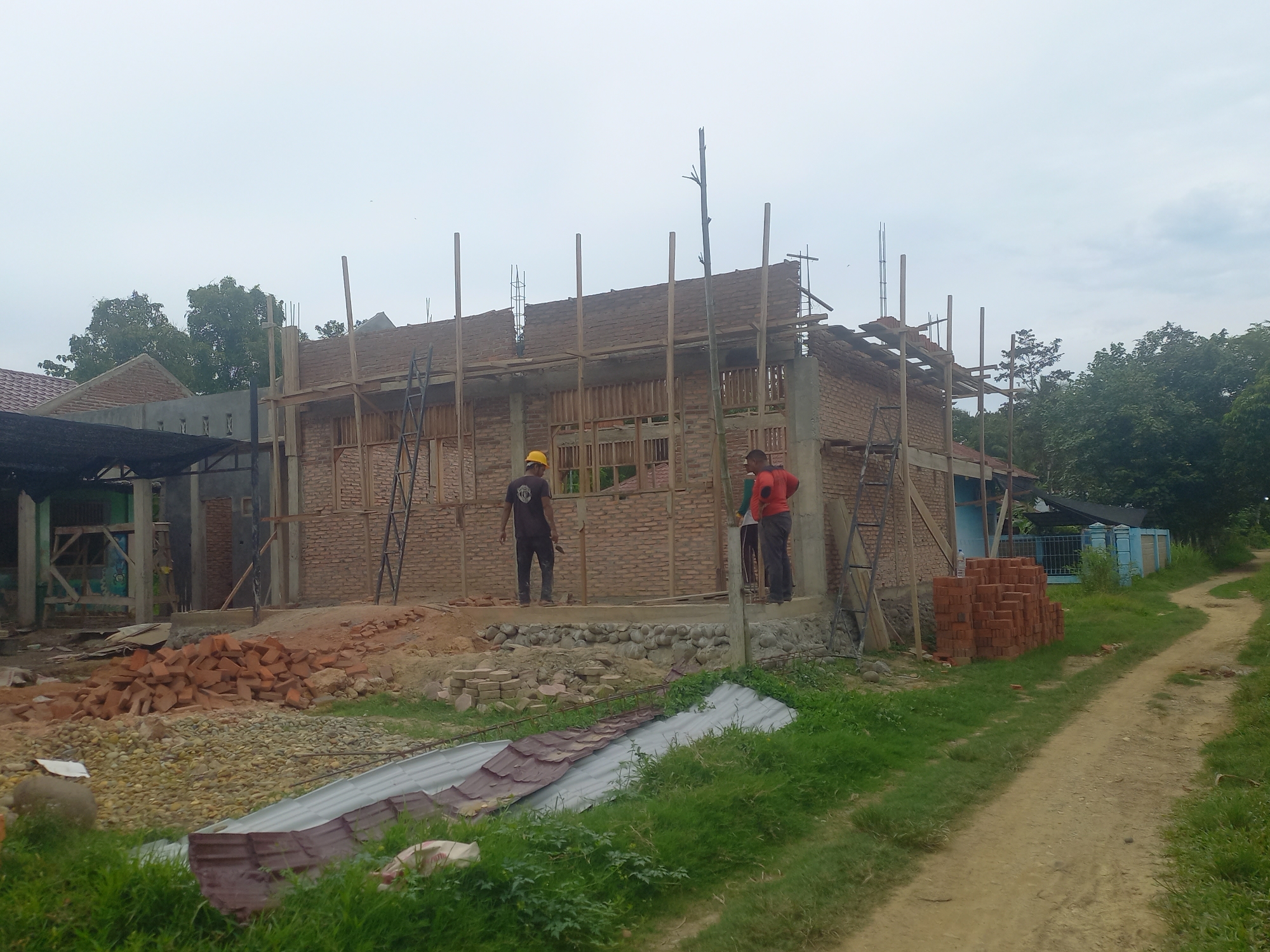 The image depicts an outdoor scene of construction in Gampongblang, Indonesia. A brick building is under construction, with wooden scaffolding supporting the roof structure. Two construction workers are visible on the partially built structure. One worker, wearing a yellow hard hat and a dark t-shirt, stands on the lower level. The second worker, in an orange t-shirt, is on an upper level, facing away from the camera. A pile of bricks is stacked on the right side of the frame, near a dirt path that winds through the grassy environment. In the background, there are trees and the roofs of other buildings, suggesting a semi-rural or suburban setting. The sky is overcast, indicating a cloudy day.