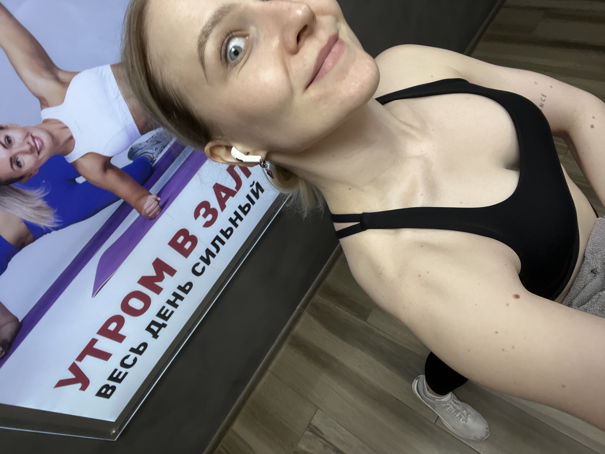 A woman is taking a selfie indoors, likely in a gym or fitness center in Khimki, Russia. She is positioned at an angle, looking towards the camera with a wide-eyed, slightly playful expression and a subtle smile. She has blonde hair pulled back, and is wearing a black sports bra, with hints of dark leggings or pants and grey sneakers visible. A white wireless earbud is visible in her left ear. On her left upper arm, a small tattoo with the partial text "NCE" can be seen, along with several moles.
Behind her, a brightly lit advertisement or poster features another smiling woman in a white sports top and blue leggings, posing actively. The poster's text is in Russian and reads: "УТРОМ В ЗАЛ" (UTROM V ZAL), which translates to "Morning in the gym," followed by "ВЕСЬ ДЕНЬ СИЛЬНЫЙ" (VES DEN SILNYY), meaning "Strong all day." The floor is made of a light brown wood or laminate. The overall setting suggests an active and health-oriented environment, possibly indicating a morning workout.