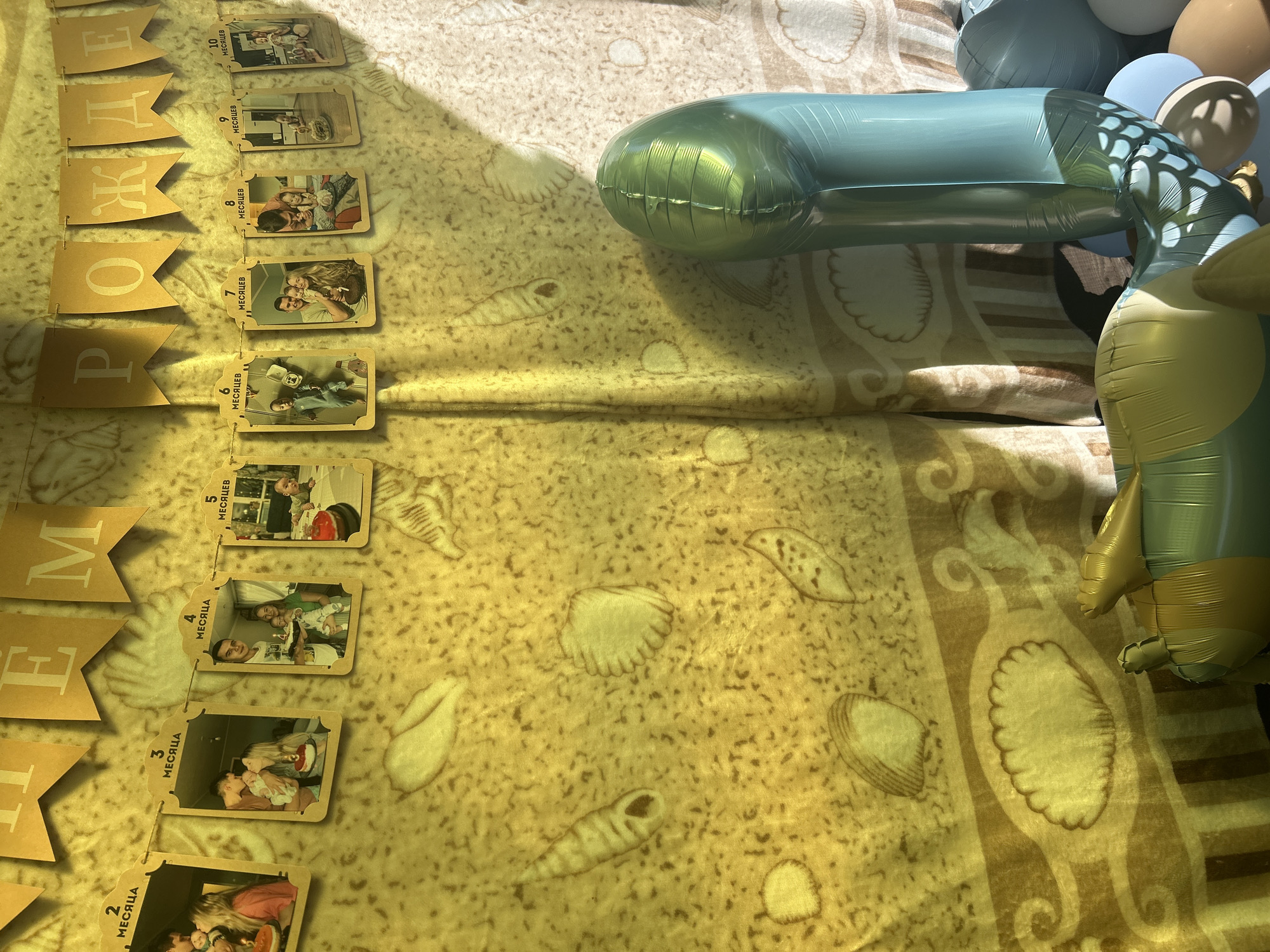 The image depicts a festive scene, likely a birthday celebration for a child, with a backdrop of a yellow banner displaying the words "ПРИ-РОЖДЕНИЕ" (birth) and a string of photos from birth up to 10 months. The photos are small, framed tags, each with a numerical label in Russian indicating the month. The backdrop itself has a sandy texture with imprints of seashells.
In the foreground, to the right, is a cluster of balloons, predominantly light blue and teal, with one large, elongated teal balloon forming the shape of the number '1'. There are also some brown and white balloons. Partially visible in the lower left is a yellow and green inflatable toy, possibly a sea creature like a turtle. The lighting suggests an indoor setting with natural light casting shadows from the right.
This scene is likely located in Lyamino, Russia, given the presence of Russian text on the banner and photo tags, and it appears to be a decorated room prepared for a child's birthday party. There are no identifiable people actively engaged in the frame; the focus is on the decorations and celebratory items.