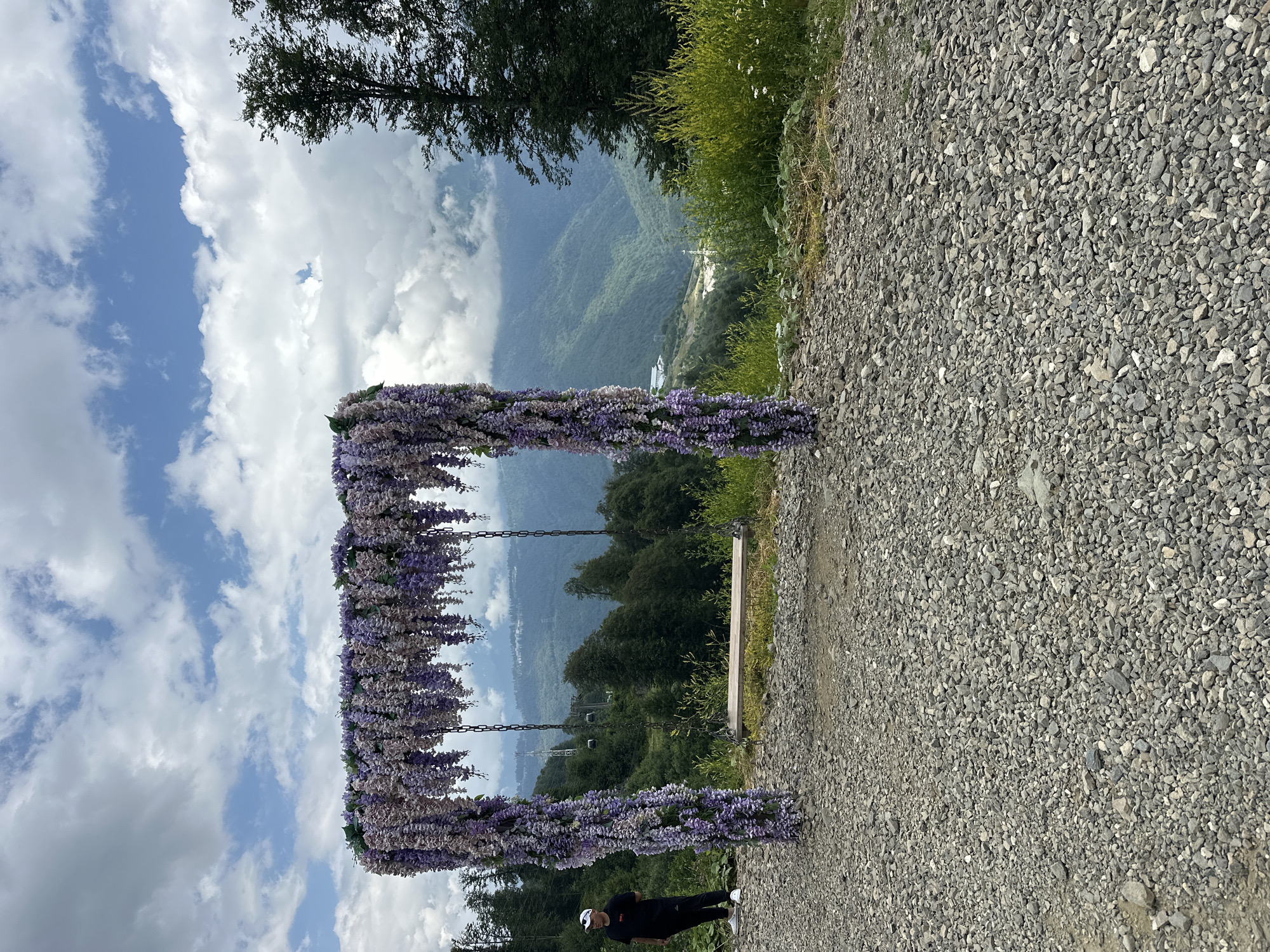 A person stands to the left of a decorative swing adorned with purple flowers. The swing has a wooden seat and is suspended by chains from a floral archway. The background features a lush, green mountainous landscape under a partly cloudy sky. The ground in the foreground is covered in gravel. The scene is set in Estosadok, Russia.