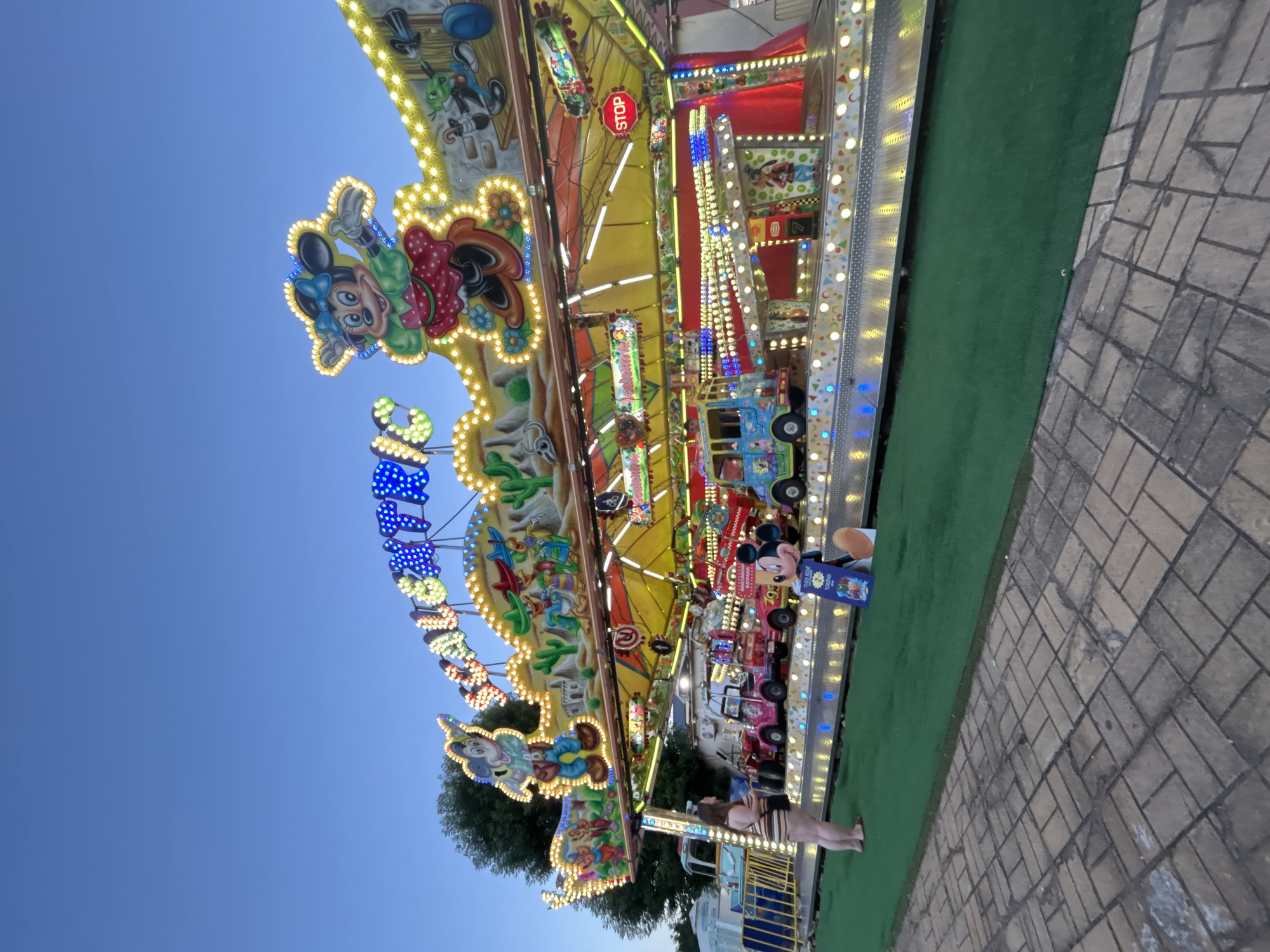 A brightly lit amusement park ride, themed with cartoon characters and a Western motif, is captured at dusk. The ride features multiple levels adorned with flashing lights and animated figures, including Mickey Mouse and Minnie Mouse. Several colorful, car-shaped gondolas are visible on the lower level. In the foreground, a young woman stands on a paved area, looking towards the ride. A life-sized cardboard cutout of Mickey Mouse holding a sign reading "THIS RIDE 2 TOKENS" is positioned on the green, artificial turf in front of the ride. The sky is a clear, deep blue, suggesting the early evening hours. The environment appears to be an outdoor amusement park or carnival.