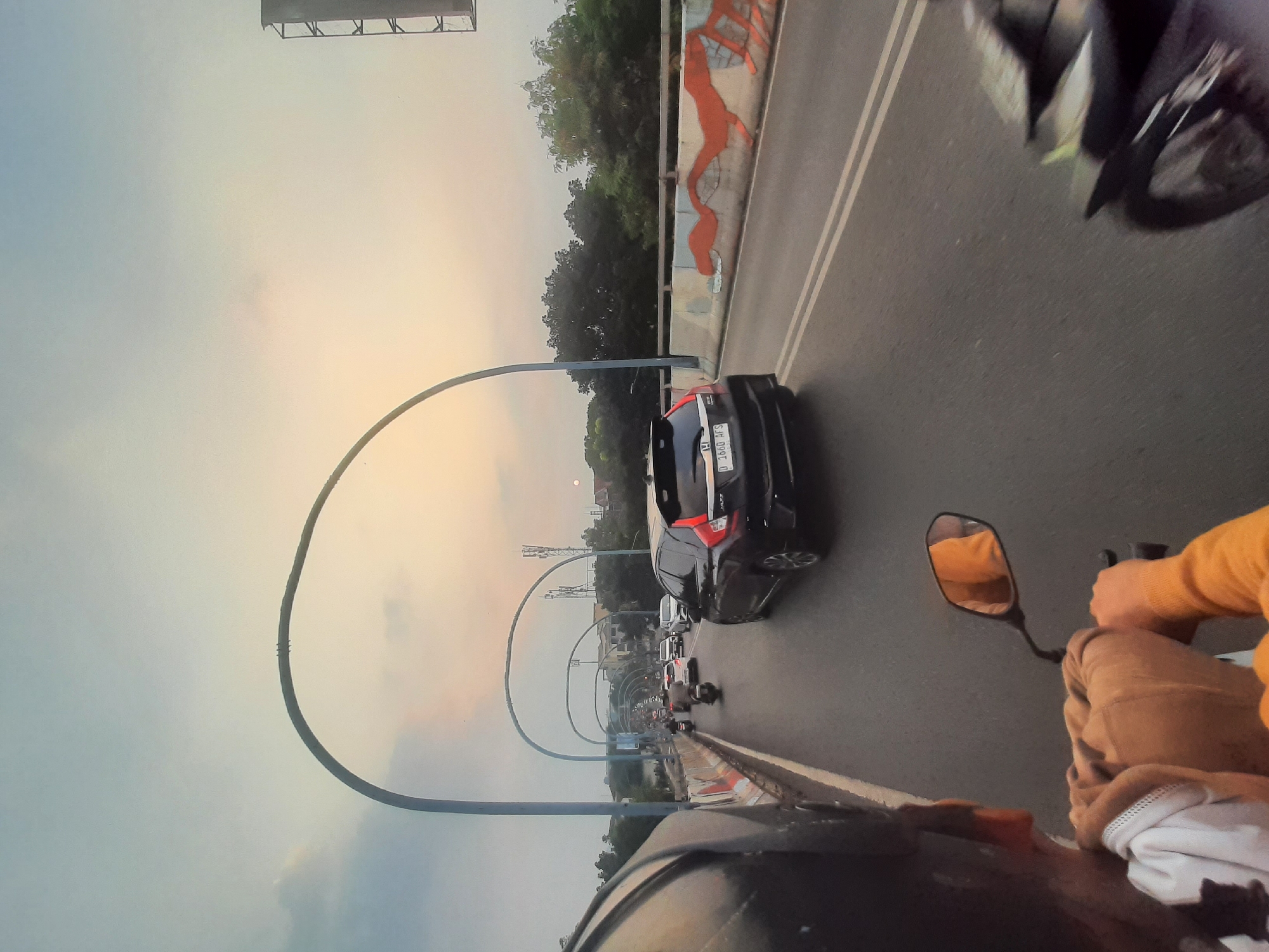 The image depicts a scene of traffic congestion on an elevated road, likely a highway overpass, at dusk. The perspective is from the back of a motorcycle, looking forward. A person wearing a helmet and a tan hoodie is visible on the motorcycle, holding onto handlebars. Another motorcycle is partially visible on the right.
In the road ahead, several vehicles are present, including a black Honda Jazz, which has a license plate that reads "D 1660 AFS". Other cars and motorcycles are also visible in the traffic flow. The road has double white lines indicating lanes.
The environment includes a concrete barrier along the side of the road, decorated with an orange, serpentine mural. Tall, arched structures, possibly lighting gantries or part of the road infrastructure, span over the highway at intervals. Beyond the road, trees and buildings are visible, suggesting an urban setting. The sky is overcast with a faint orange glow near the horizon, indicative of sunset.
Given the license plate format "D 1660 AFS", the 'D' prefix is a strong indicator that the vehicle is registered in the West Java region of Indonesia, which includes Bandung City. The presence of such elevated roads and traffic conditions is consistent with urban areas like Bandung.