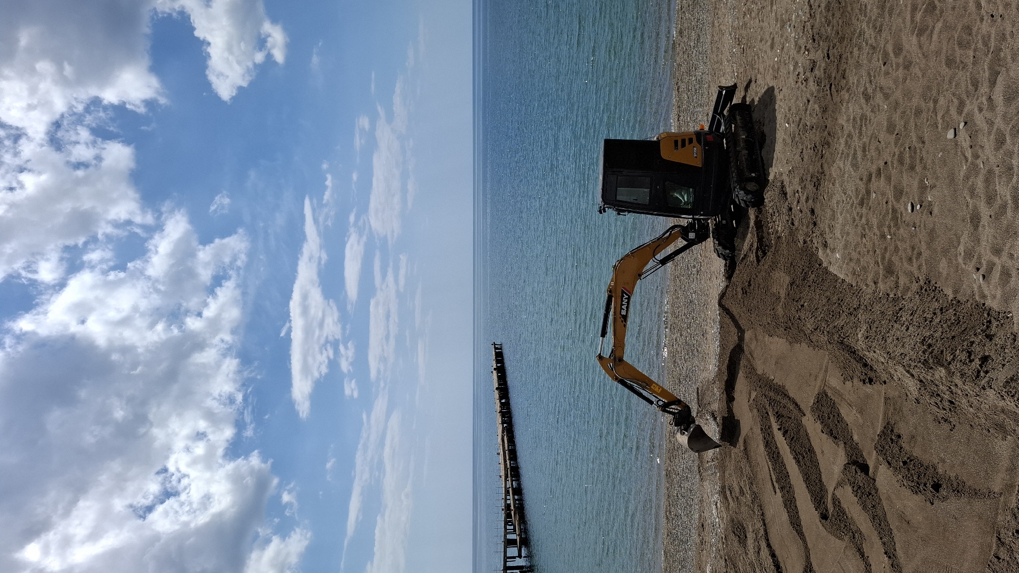 A beach scene in Unknown, Unknown is depicted on a sunny day with a blue sky scattered with cumulus clouds. The sea is visible on the left side of the frame, with a weathered pier stretching into the water. A yellow and black excavator is positioned on the sandy beach, actively digging or moving sand near the water's edge.