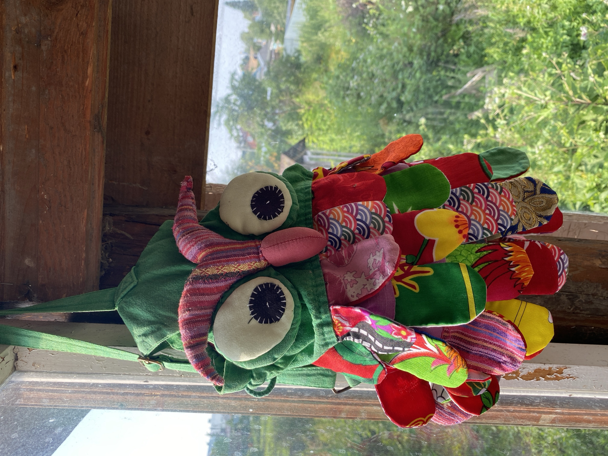 A stuffed, owl-shaped decorative object is hanging from a wooden beam near a window. The owl is primarily green with large, light-colored eyes and a pink beak. It is adorned with various colorful fabric patches in patterns of stripes, scales, and floral designs, which form its "feathers." The object is suspended by a green fabric strap. The window shows a view of trees and buildings, suggesting a rural or suburban setting. The overall lighting indicates it is daytime. The location is identified as Zelenodol'sk, Russia, though no specific details within the image confirm this.