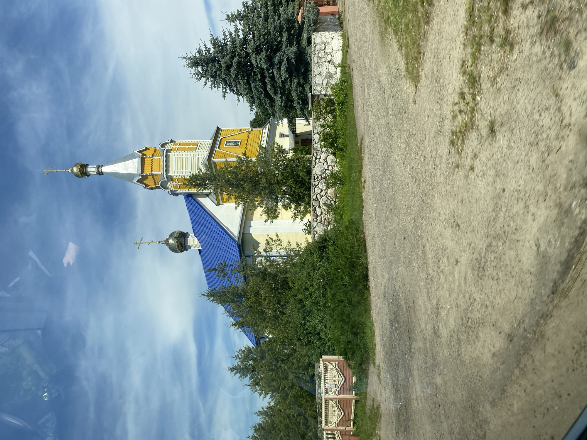 The image shows a yellow wooden Orthodox church with a blue roof and a silver and gold dome. It is located in the village of Verkhneye (Верхнее) within the Volchynsky Selsoviet (Воўчынскі сельскі Савет) in Belarus. The church is surrounded by trees and a stone wall. In the foreground, there is a dirt road and a decorative fence. The sky is blue with some clouds. The scene appears to be captured from a moving vehicle, as indicated by reflections on the glass in the upper part of the image.