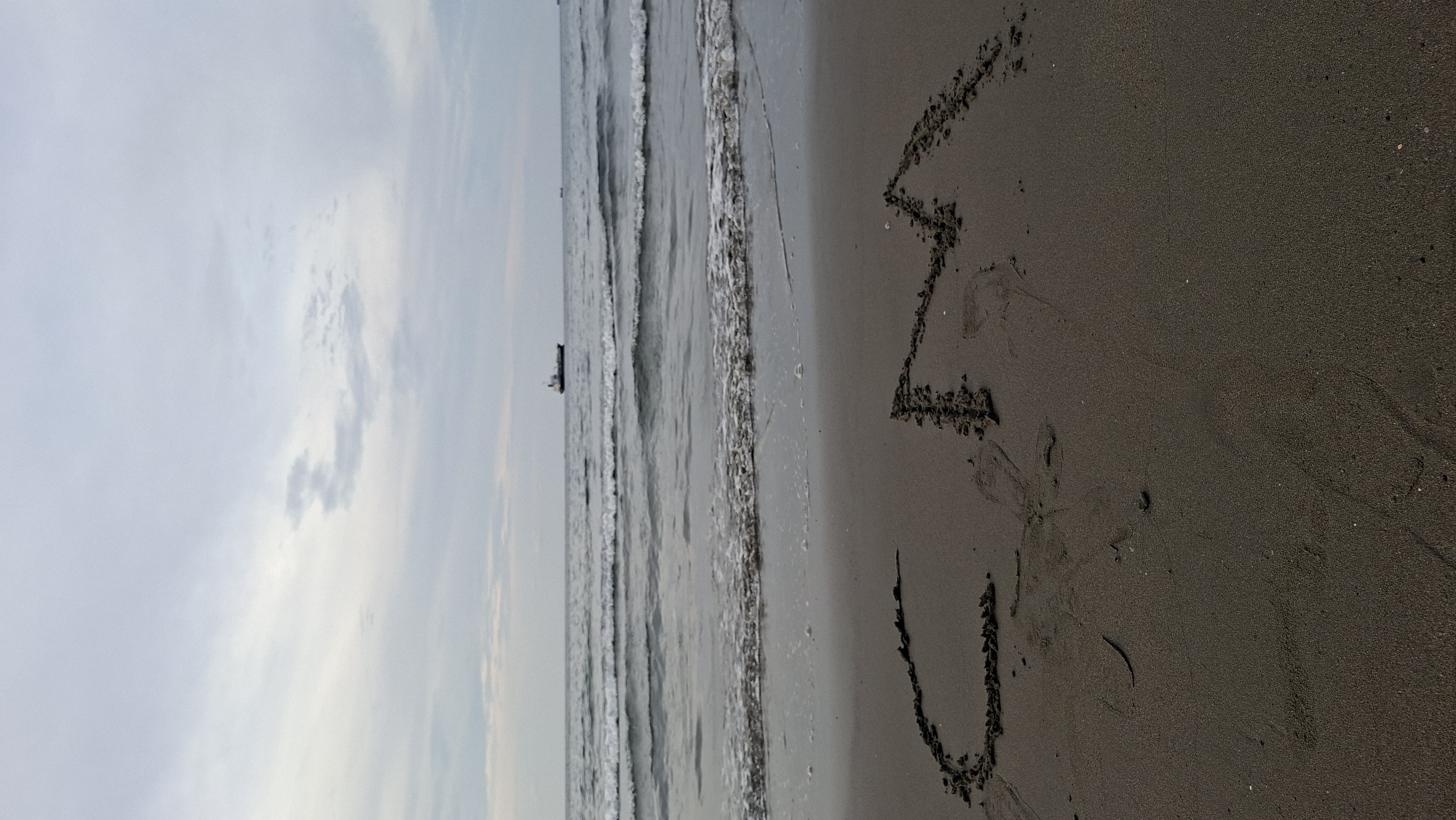 A wide shot captures a beach scene with the letters "CM" drawn in the sand in the foreground. The ocean with gentle waves stretches out behind the beach, and a ship is visible on the horizon under a cloudy sky. The location is the beach in Lhokseumawe, Indonesia.