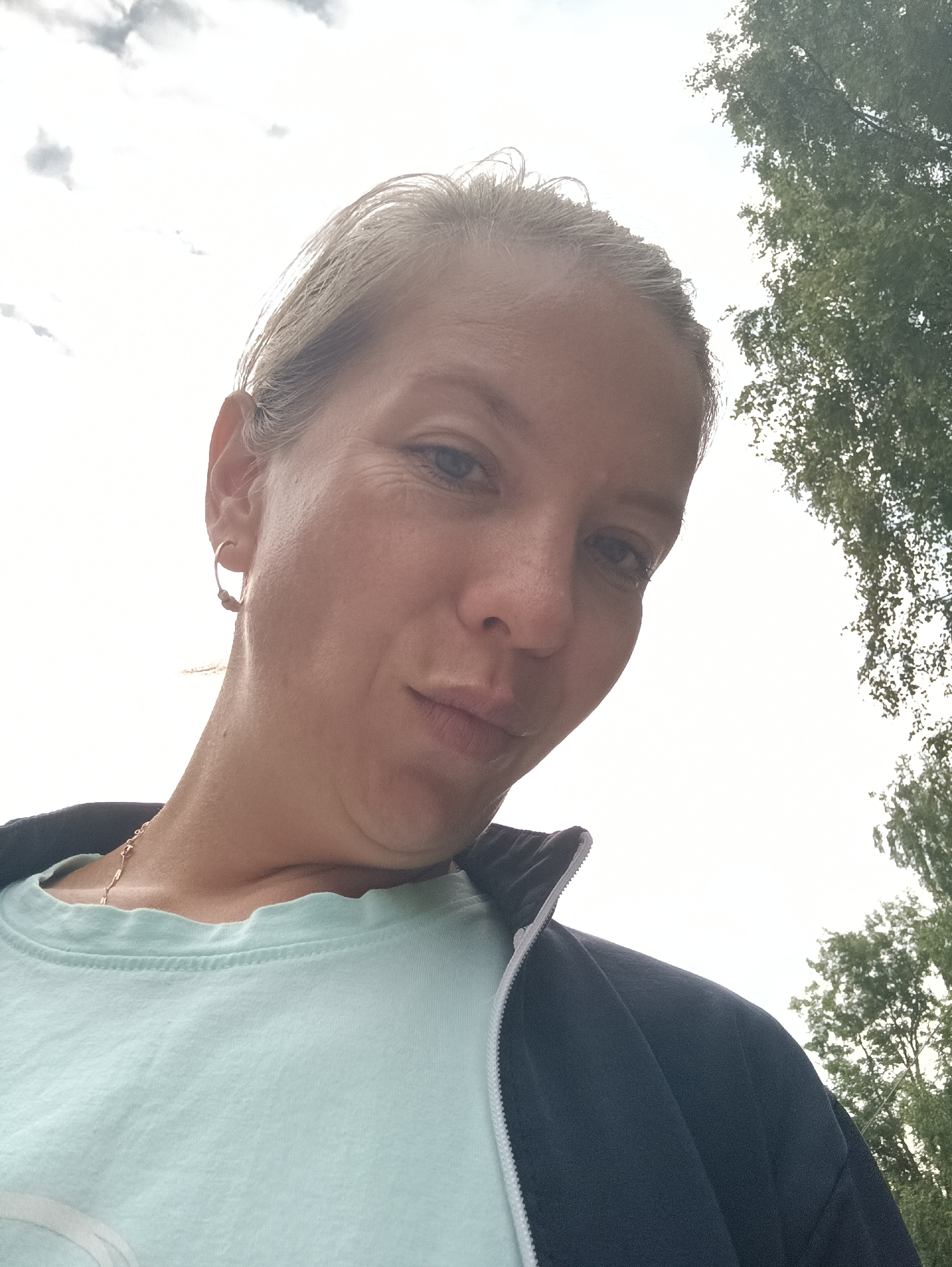A low-angle, medium close-up shot shows a fair-skinned woman with blonde hair pulled back. She is wearing a light blue t-shirt and a dark blue zip-up jacket. A thin gold chain necklace is visible. The woman is looking slightly upward and to the left, with her lips pursed. Her left earlobe has a small hoop earring. The background is a bright sky with some clouds and the upper branches of a green tree. The image appears to have been taken outdoors in Chaikovsky, Russia, likely in a park or residential area.