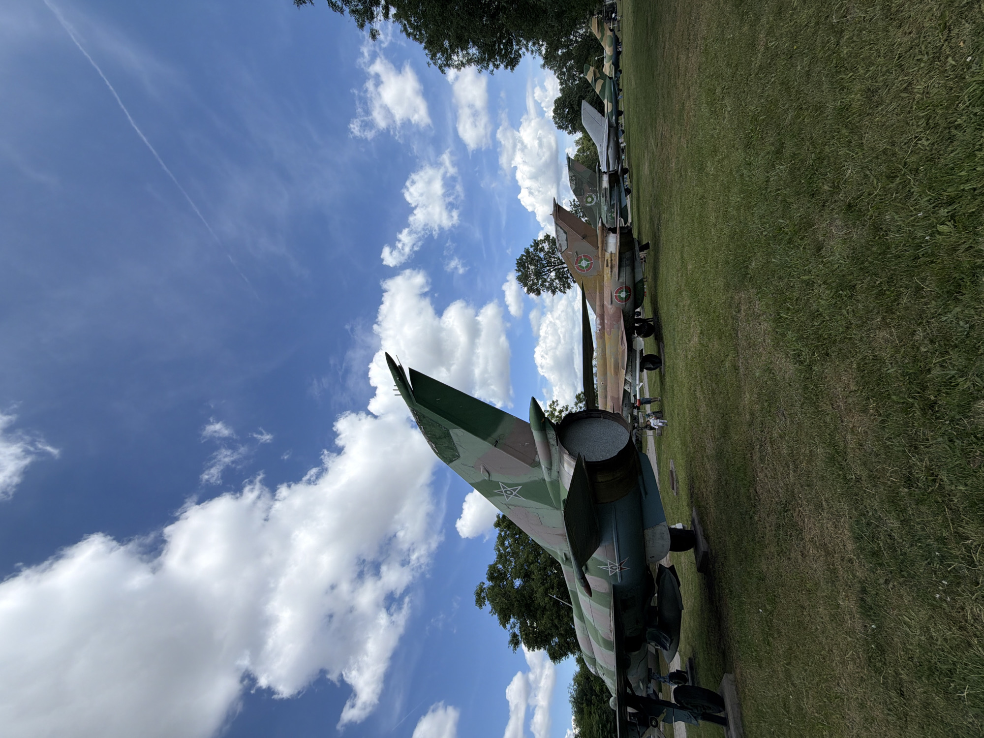 The image shows a lineup of parked fighter jets on a grassy field under a cloudy blue sky. The jets are camouflage-painted and appear to be on display. In the foreground, the tail of a green and brown camouflage jet is prominent. In the background, several more jets are visible in a line, along with trees on the right side of the frame. A few people are scattered on the field near the jets. This scene is consistent with an outdoor museum or exhibition, such as the Museum of Aviation in Krumovo, near Plovdiv, Bulgaria, or an airfield display. The presence of multiple Soviet-era fighter jets suggests a military history context. The lighting and shadows indicate it is daytime.