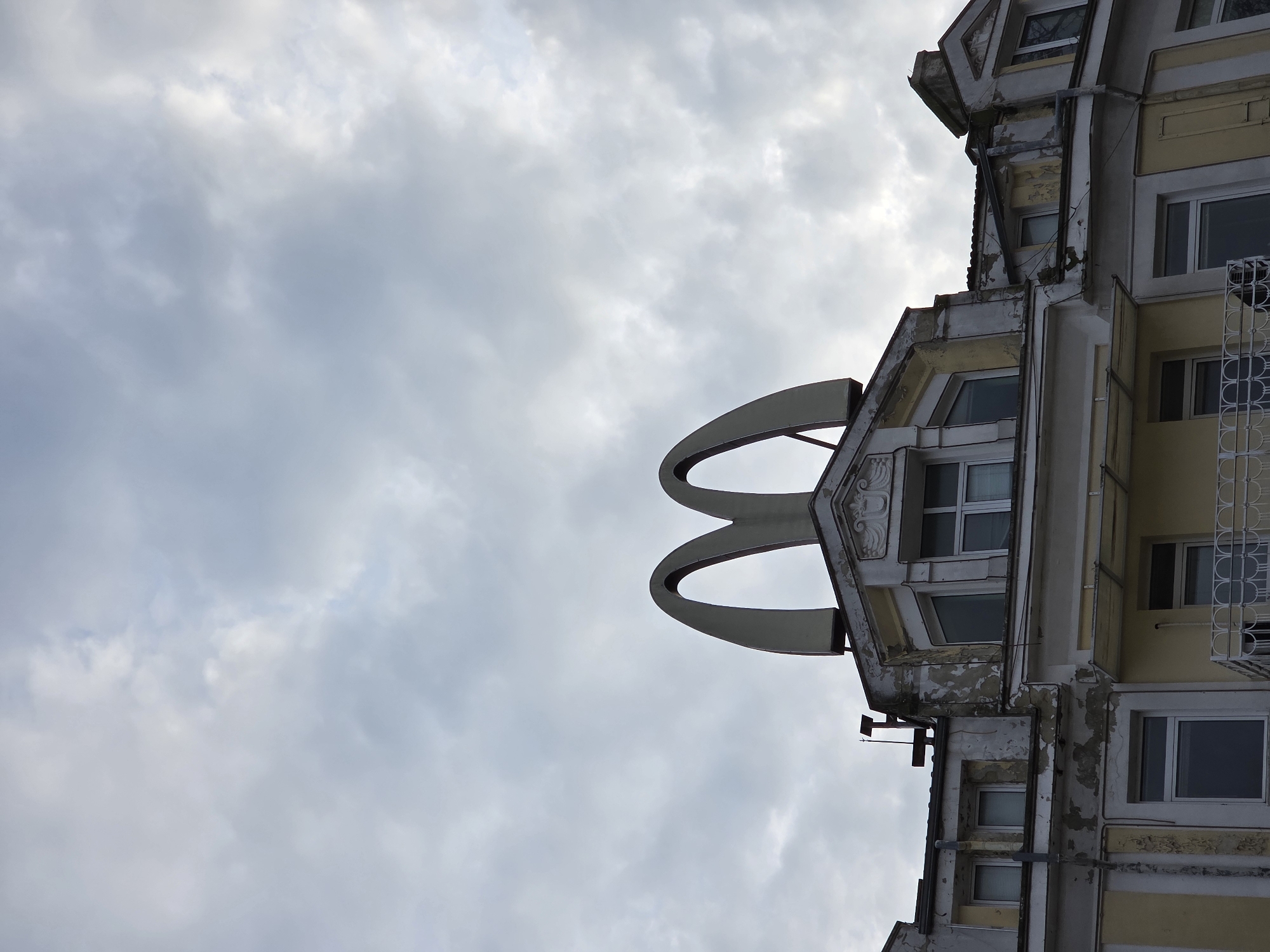 An unusual building facade, likely once housing a McDonald's restaurant, is seen in Varna, Bulgaria. The iconic golden arches are attached to a multi-story building, positioned on the top. The building's facade shows signs of wear and tear, including peeling paint. The sky above is overcast with clouds.
