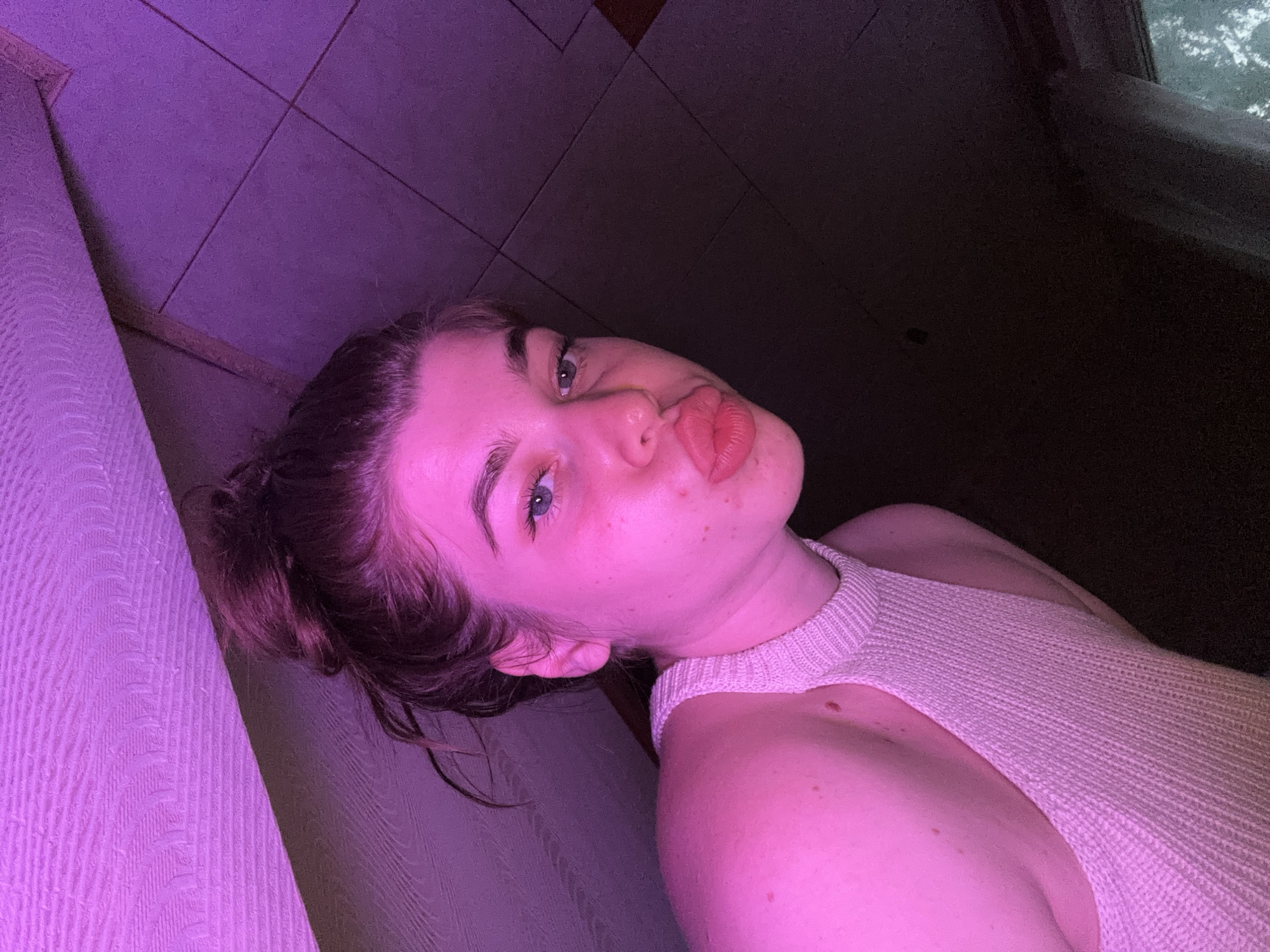 The media file is a portrait of a young person, oriented sideways. The person has light skin, light blue eyes, and dark hair pulled up in a bun. They are wearing a light-colored knitted top with a halter neckline. The person's face is turned towards the camera, with their lips puckered in a kissing gesture. A mole is visible on their left shoulder. The background is dark, with hints of a tiled surface illuminated by purple lighting on the left side of the frame. The overall mood is playful and intimate, possibly taken in a dimly lit indoor setting in Мещёры, Russia. No visible text is present.