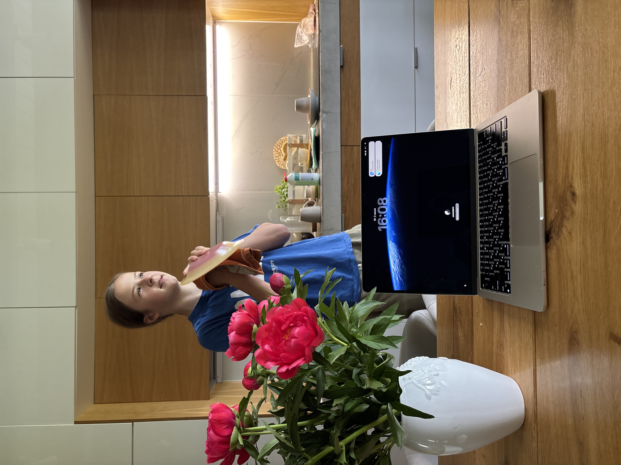 This image, captured in Chaikovsky, Russia, portrays a domestic scene indoors, likely a kitchen or dining area, during the late afternoon. A young child, with light brown hair, wearing a blue t-shirt with partially visible text "THE CITY", is positioned on the left side of the frame. She holds what appears to be a book or tablet, resting on an orange napkin, and looks upwards and slightly to the right with a calm, contemplative expression.
On a wooden table in the foreground, a prominent bouquet of vibrant pink/red peonies is displayed in a white, textured vase. To the right of the flowers, an open silver laptop, resembling a MacBook, rests on the table. Its screen shows a dark wallpaper, possibly a planet or space scene, with the digital time "16:08" prominently displayed, indicating it is 4:08 PM. Notification icons are visible at the top of the screen, and smaller text shows "BT 2 home" and "ВТ-1_Ростелеком", likely network names.
The background features modern cabinetry: white, flat-panel cabinets are on the left, while wooden cabinets line the wall above a countertop on the right. The countertop holds various kitchen items including mugs, cups, a straw hat, and a bottle of cleaner, illuminated by under-cabinet lighting. The overall impression is a quiet, everyday moment within a well-appointed home.