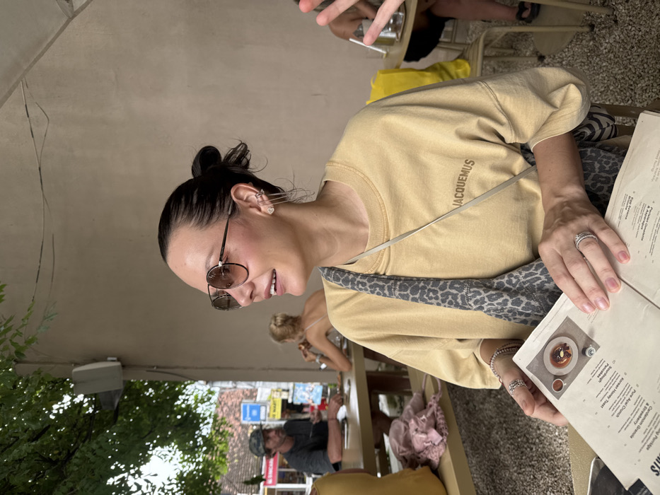 The image captures a woman seated at an outdoor cafe in Uluwatu, Indonesia, engaged in perusing a menu. The woman, positioned in the mid-left foreground, has dark hair pulled back, wears brown-tinted aviator sunglasses, and smiles towards the right side of the frame. Her left ear features multiple piercings, including a stud and a cascade of thin chains. She is dressed in a mustard yellow crewneck sweatshirt with "JACQUEMUS" printed in brown on the chest. A grey and black leopard print cross-body bag strap is visible over her right shoulder. Her left hand, adorned with multiple rings, holds an open menu displaying food items such as "Sourdough," "Korean Honey Toast," "Pancakes," and images of plated food, including a dessert and a coffee cup.
The background reveals a street scene to the far left, featuring a light-colored building with a red roof, storefronts (one partially visible as "MINI MART"), and a blue and white road sign. Green foliage from an overhead tree canopy is visible. A man with a beard and a cap, wearing a dark t-shirt, is seated at a light brown wooden table further down the street, facing left. Another individual with blonde hair is partially visible at a table behind him. The immediate cafe environment includes light brown walls, a white speaker mounted on the wall to the woman's left, and a ground surface composed of light-colored gravel or crushed stone. Various light brown wooden tables and chairs are scattered throughout the cafe area. To the far right, a blurred yellow bag and a person's hand are discernible at another table.