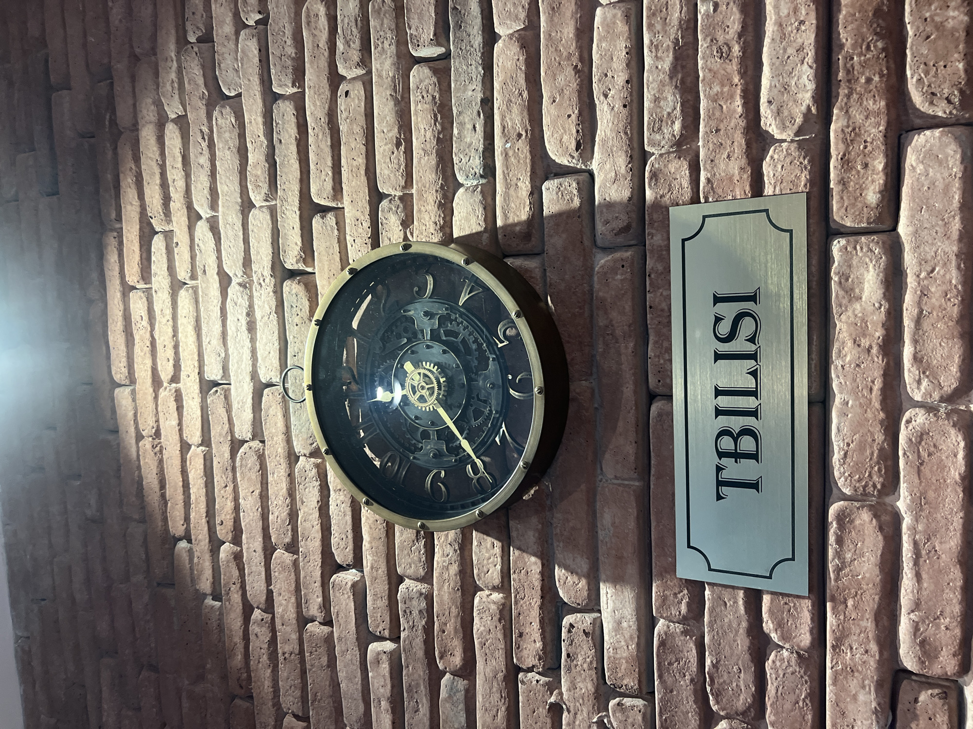 An ornate, vintage-style clock with a brass frame and a dark face hangs on a rustic brick wall. The clock features Roman numerals and visible internal gears, suggesting a mechanical movement. Below the clock, a rectangular metal plaque with a decorative border displays the word "TBILISI" in bold, serif font. The brick wall appears weathered, with variations in color and texture, indicating an interior setting. The lighting casts shadows on the wall, highlighting the brickwork. The scene is static and devoid of human presence.