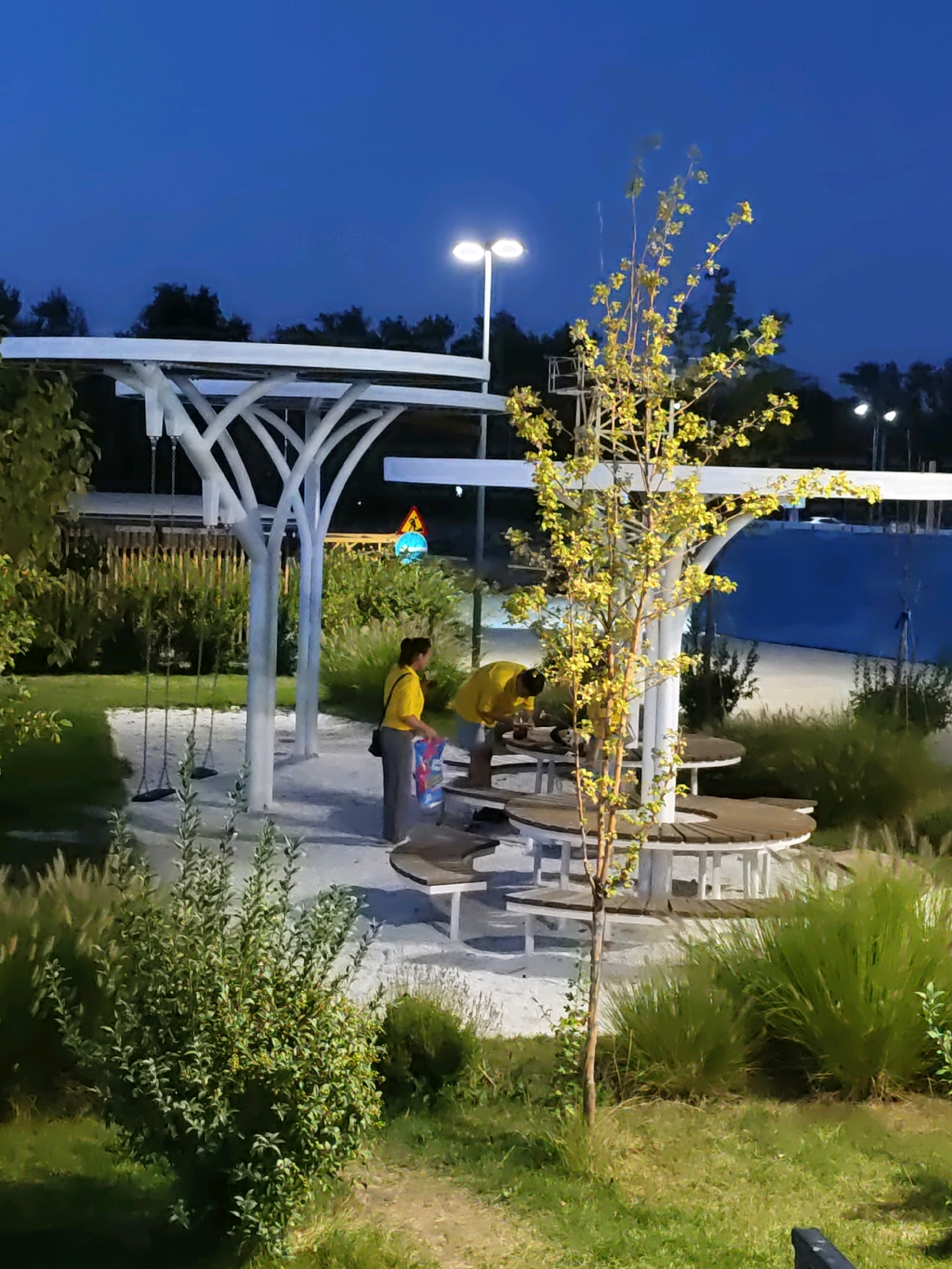 The image depicts an outdoor seating area at dusk, with a young tree in the foreground and a covered pavilion with picnic tables behind it. Two individuals, both wearing yellow shirts, are interacting at one of the tables. One person is kneeling, seemingly inspecting something on the table, while the other stands nearby. Behind the pavilion, there is a body of water, possibly a pool or a river, and a blue structure. The area is illuminated by streetlights and the remaining natural light. In the distance, trees and a traffic sign are visible. The overall setting appears to be a public park or recreational space.
