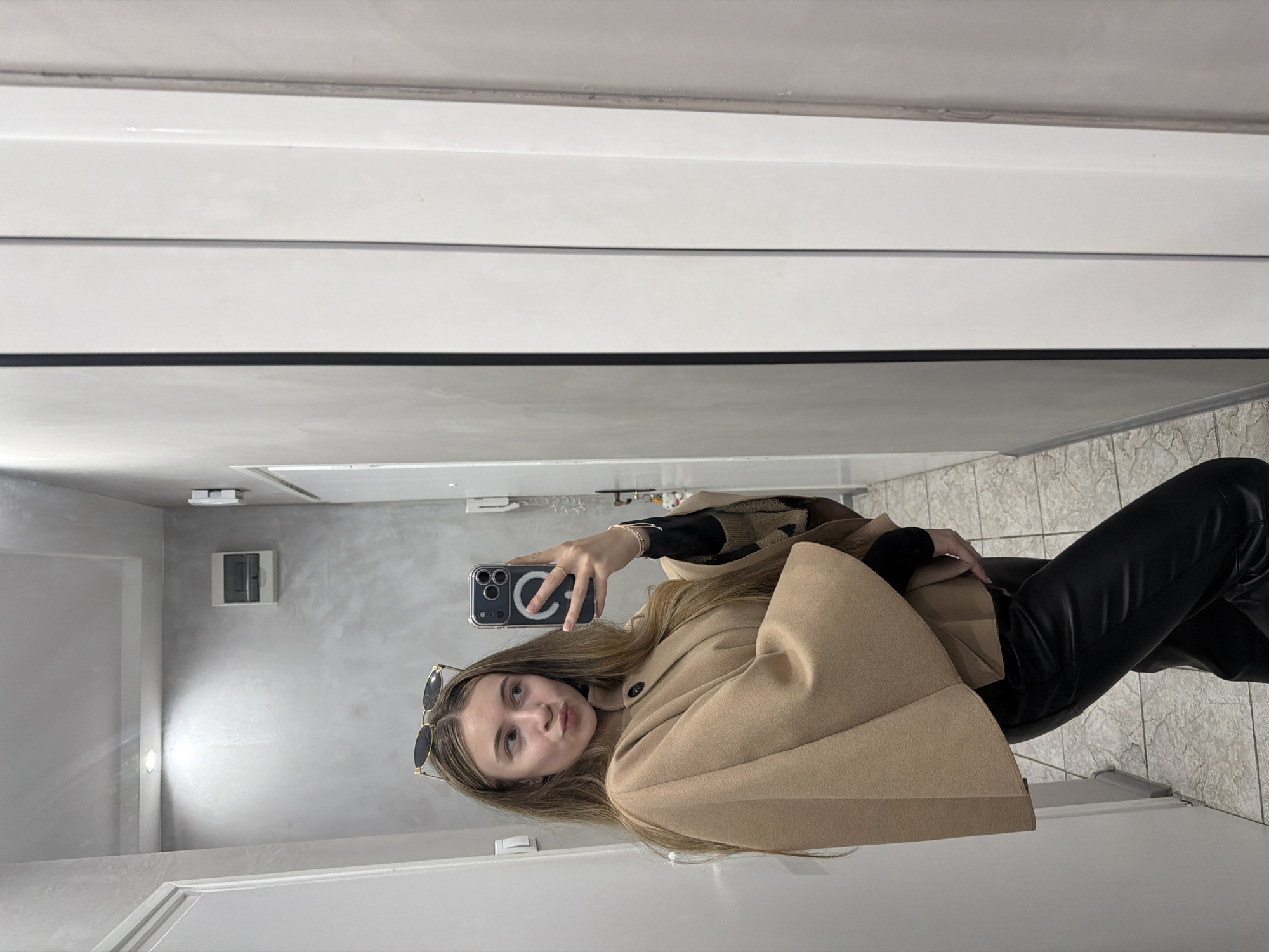 A woman is taking a selfie in a restroom. She is wearing a light brown coat, black pants, and has sunglasses perched on her head. The restroom has white tiled walls and floor. A gray wall can be seen on the left side of the image, where a light fixture is visible. There's also a gray electrical box on the wall. The location is not identifiable as Plovdiv, Bulgaria from the image.