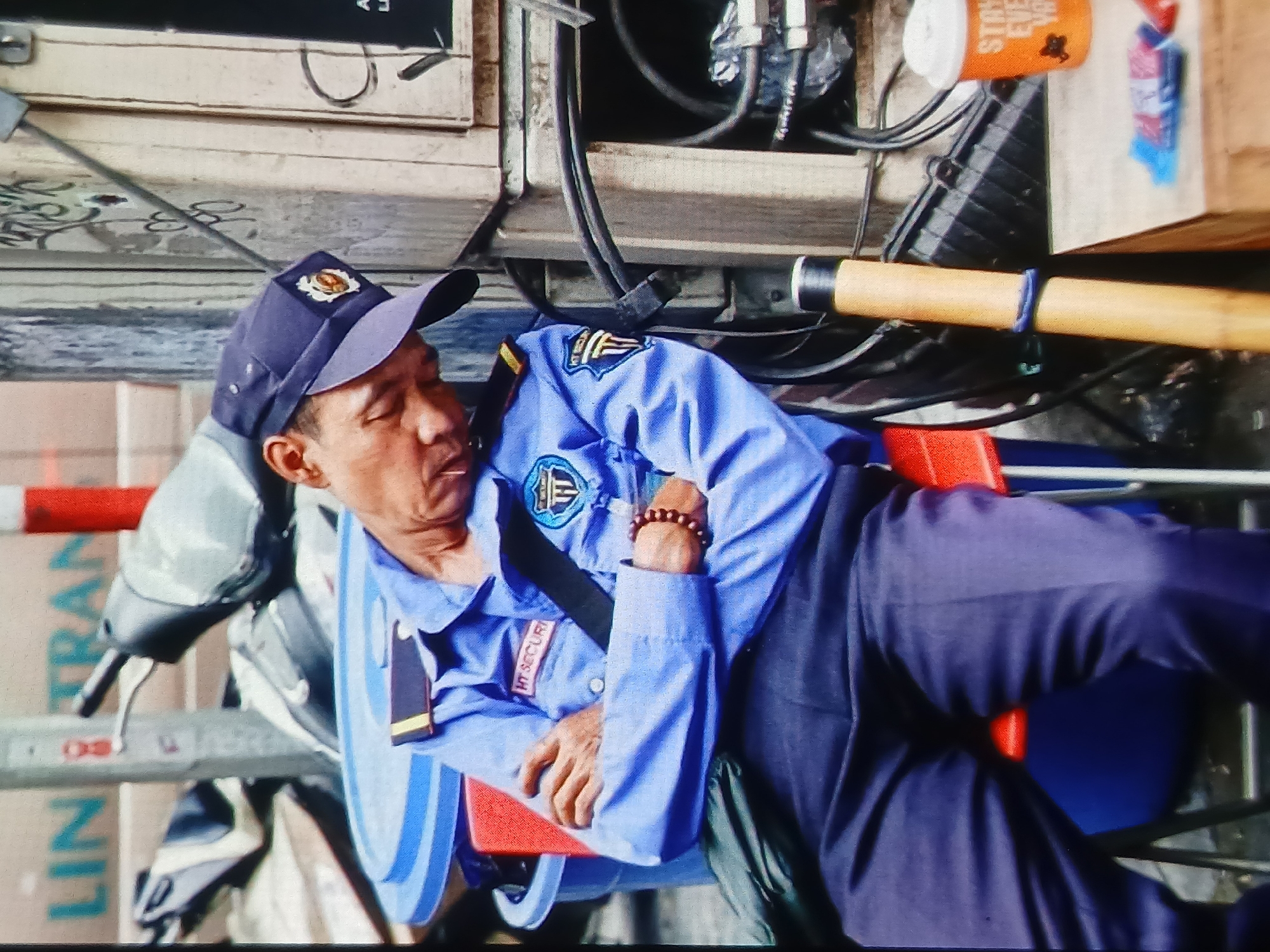 A man in a blue security guard uniform, complete with a cap, is asleep with his arms crossed. He is seated on a red chair. The uniform features several patches, including one on the left breast that reads "HT SECURITY" and another on the sleeve with a crest. He is wearing a dark-colored bracelet on his right wrist.

The background suggests an urban environment. To the left of the man, a blue bin is visible, partially obscured by a green bag. In the middle ground, motorbikes are parked. The man appears to be in a street or alleyway, with electrical conduits and wiring visible on a vertical surface to his right. An orange cup with text, possibly an advertisement, sits on a wooden surface in the lower right corner, next to some packets of what appear to be candies. The lighting suggests it is daytime. The overall scene depicts a moment of rest or inactivity by a security guard.

The location is identified as Setia Bakti, Indonesia.