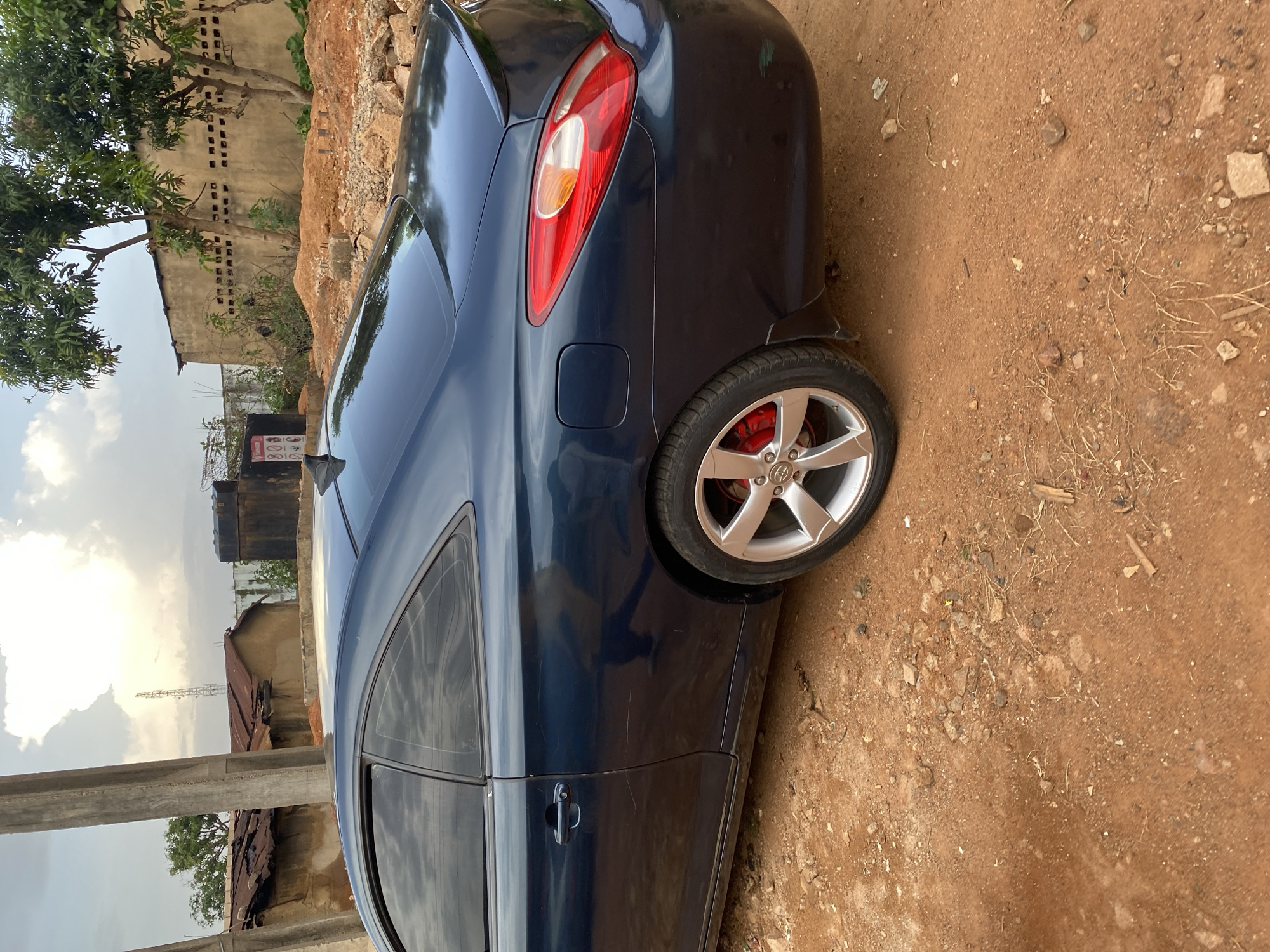 In Bukuru, Nigeria, a dark blue coupe is parked on a dirt road. The car is viewed from the rear-right side, with its driver's side door, rear fender, taillight, and spoiler clearly visible. The vehicle has silver rims, red brake calipers, and tinted windows. The surrounding environment includes a large concrete pillar on the left, a corrugated metal roof on a building behind the car, and a wall with several small windows in the background. Trees with green leaves are also present, and the ground is covered with reddish-brown dirt, small rocks, and scattered debris. The sky is cloudy with patches of sunlight.