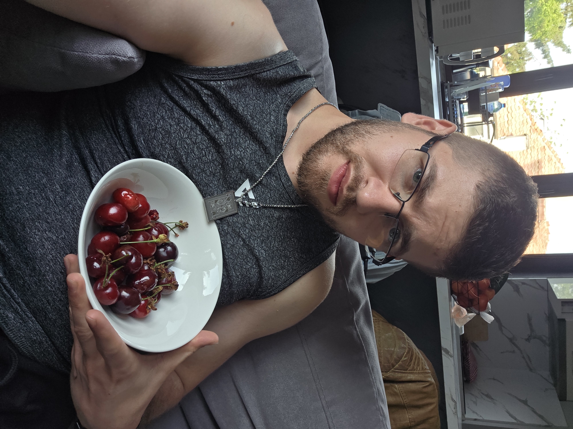 A person is sitting on a gray couch, holding a white bowl filled with red cherries. The person is wearing a gray tank top and glasses, and has a beard and a silver necklace. In the background, there's a window with buildings visible outside, suggesting an urban setting. Considering the request for location specificity, and without visual confirmation, the image may have been taken indoors in Yagodovo, Bulgaria. The scene appears casual and domestic, with the person seemingly offering or enjoying the cherries.