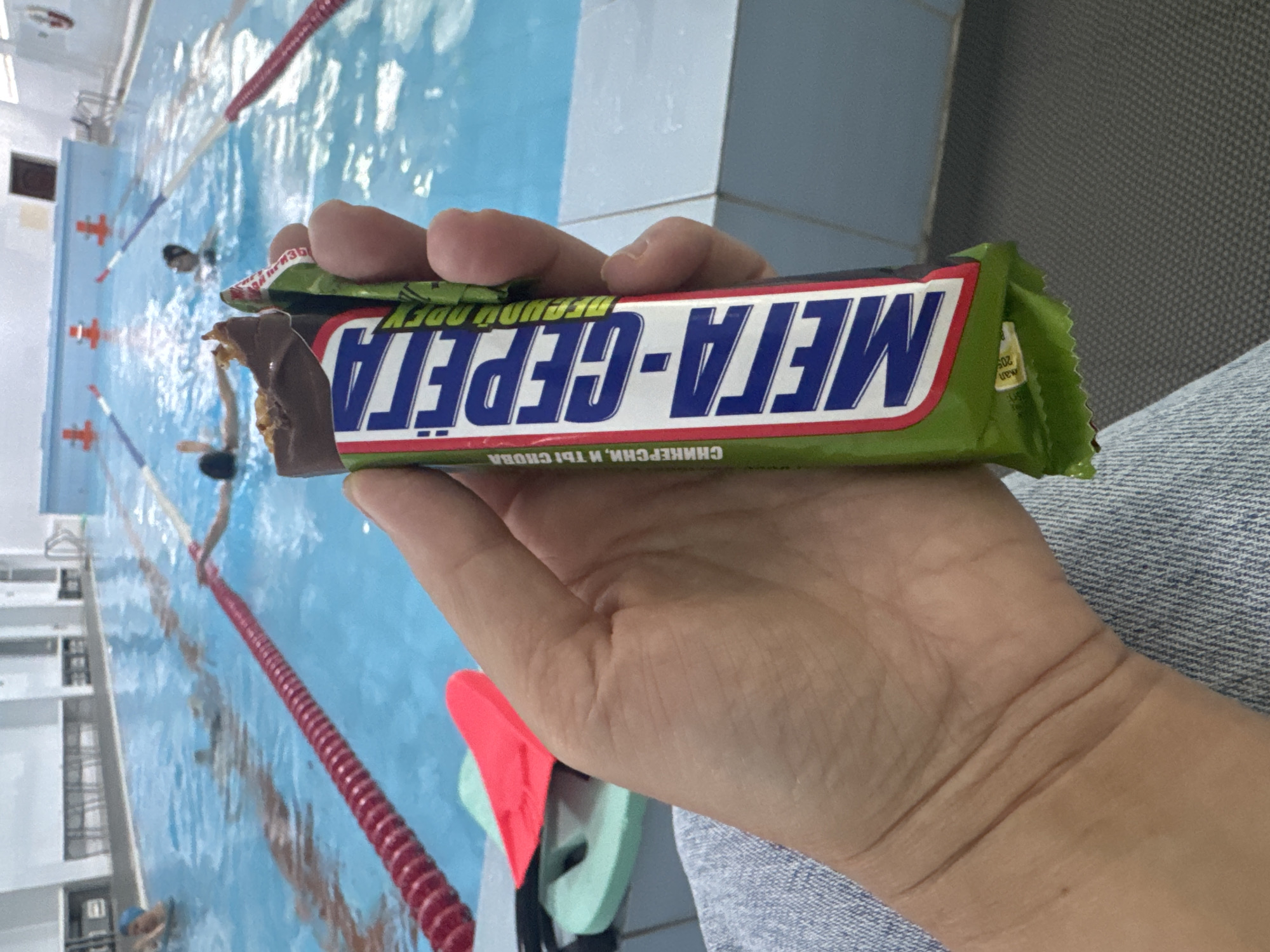 The image captures a close-up of a person's hand holding a partially eaten chocolate bar, set against the backdrop of an indoor swimming pool. The hand is visible in the lower center, with fingers wrapped around the chocolate bar. The person appears to be wearing light blue denim or similar fabric, visible on the right.
The chocolate bar wrapper is predominantly green with a red and white border. Bold blue Cyrillic text on the wrapper reads "МЕТА-СЕРЕПА" (META-SEREPA), likely the brand name. Below this, a tagline in smaller white text on a green strip is visible: "СНИКЕРСНИ, И ТЫ СНОВА" (Snickers yourself, and you are back), a clear play on the famous Snickers slogan. Part of another text, possibly "ОБЕДУЙ" (OBEDUY - have lunch/dine), is visible on the side of the wrapper, along with small digits "205" and the word "new". The chocolate bar itself shows a brown exterior and a multi-layered interior with nougat, caramel, and peanuts where a bite has been taken.
In the background, a brightly lit swimming pool with clear blue water is visible. Red lane ropes stretch across the pool. At least three individuals are in the water, appearing to be swimming or engaging in water activities. On the pool deck, near the bottom right, swimming equipment like kickboards (one red, one light green) can be seen. The walls of the facility are white, and an overhead light source is reflected in the water.
The scene suggests a casual moment, possibly a break or post-activity snack by the pool, in an indoor setting in Birobidzhan, Russia, given the Cyrillic text on the packaging.