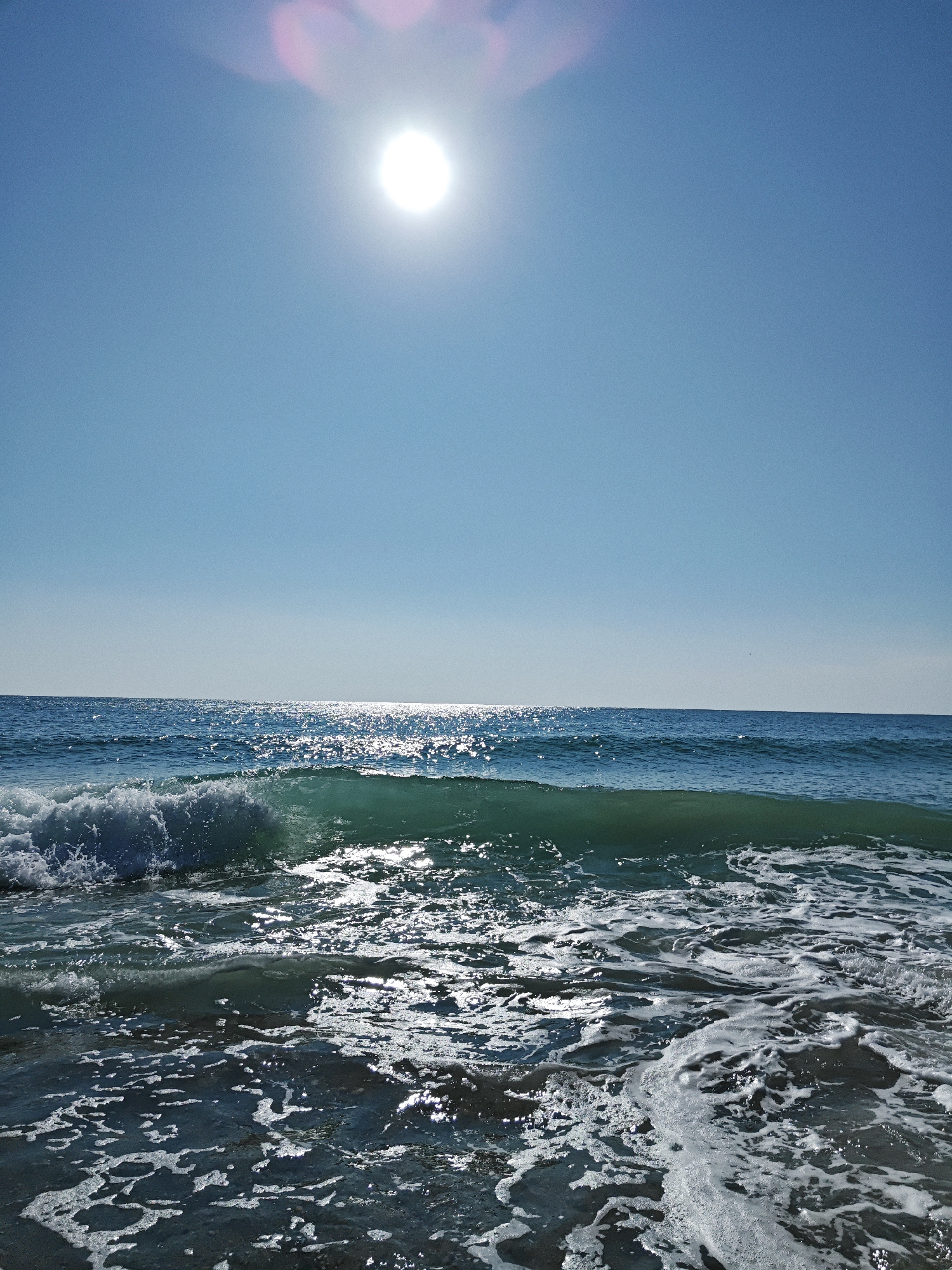 A bright sun shines in a clear blue sky above the ocean. Waves crash onto a beach, creating white foam and spray. The water is a deep blue, with sunlight glinting off its surface. The scene is peaceful and serene.