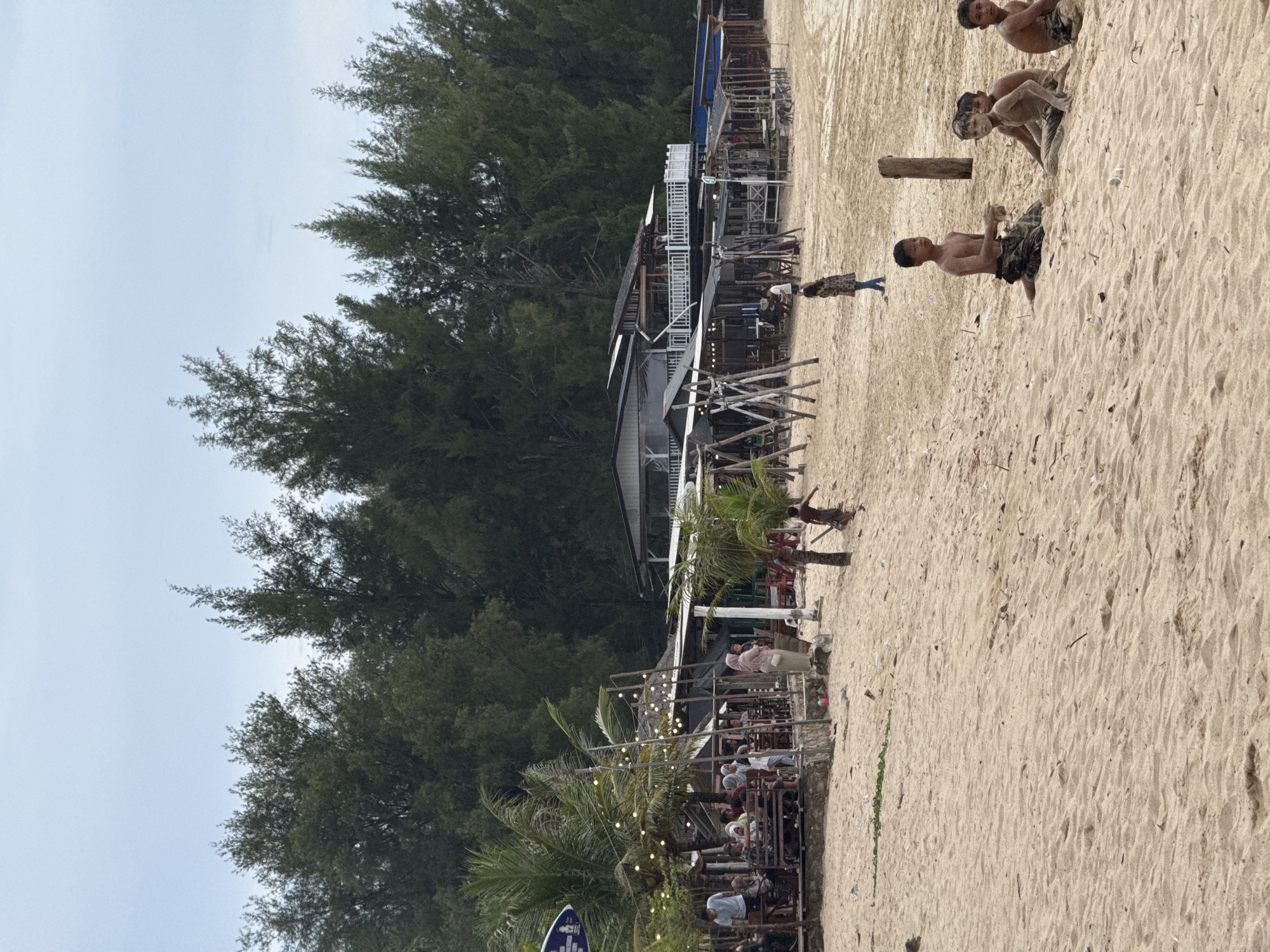 The image depicts a beachside scene in Meunasahbale, Indonesia, characterized by a sandy shore backed by dense pine trees. In the foreground, several young boys, some with sandy skin, are seated on the beach. Further back, a variety of structures line the edge of the sand, including open-air seating areas with tables and chairs, some decorated with string lights. A multi-story building with balconies is visible in the background, suggesting a restaurant or resort. People are seen sitting at tables, walking, and interacting in these shaded areas. The overall atmosphere appears to be a casual, recreational setting on a cloudy day.