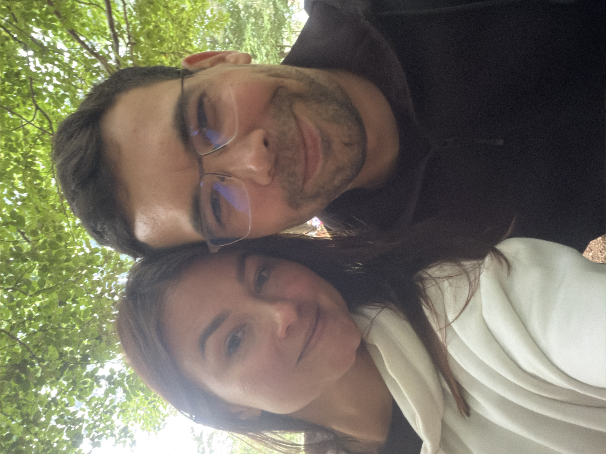 The image displays a close-up, sideways-oriented photograph of two individuals, a man and a woman, posing together outdoors.
The man is positioned on the left side of the frame (when viewed with his head upright). He has dark hair, a beard, and wears clear-framed glasses that show reflections of light. He is dressed in a dark hooded top and has a faint, pleasant smile.
The woman is situated to his right and slightly below him, with dark hair cascading over her shoulders. She is wearing a light-colored, possibly white, top or jacket, featuring soft folds, over a darker garment. She also exhibits a gentle smile.
The background consists of a vibrant green canopy of tree leaves and branches, through which bright sunlight filters, indicating a natural outdoor setting during the daytime. The lighting suggests clear weather. The overall mood appears calm and pleasant. There is no visible text in the image. This scene is consistent with the provided location context of Mali Bostur, Montenegro, a region known for its natural landscapes.