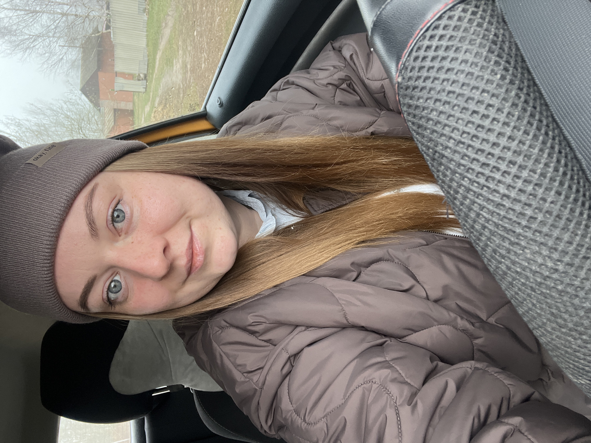 The image shows a woman with light skin, wearing a brown beanie and a brown quilted jacket, positioned inside a vehicle, likely a car. The passenger seat is visible to her right. The exterior visible through the car window shows a building and trees in Раздолье, Russia. It appears to be a foggy or overcast day.