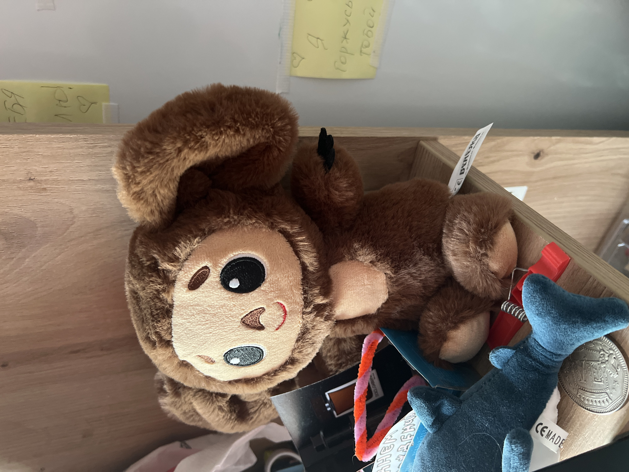 A close-up shot shows a brown plush monkey toy lying on its side in a wooden box. The monkey has a round face with large, expressive eyes, a small nose, and a smiling mouth. Its fur is soft and fluffy, and its limbs are relaxed. Other objects are visible in the box, including a small blue shark plush toy with its tail visible, a red clip, and a metal coin. There are also some colorful pipe cleaners and a white tag with text that appears to say "CE MADE." A piece of yellow paper with handwritten text is attached to the wall behind the box, and another yellow note is visible to the left. The lighting suggests an indoor setting, possibly a room or storage area. The overall scene appears to be a collection of toys and small items in a disorganized yet homey environment.
