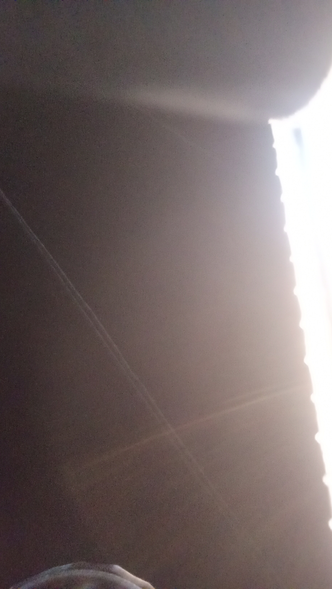 The image depicts a dimly lit environment with a bright light source emanating from the right side of the frame. Two faint, parallel streaks of light are visible across the dark expanse of the image, possibly suggesting movement or reflections. In the lower left corner, a blurry section of what appears to be a person's head or shoulder is visible, with some indication of hair or fabric. The overall impression is that of an indoor or shaded area, with the bright light creating a dramatic contrast and partially obscuring details within the scene. The image lacks any specific landmarks or context that would allow for definitive identification of its location within Kuta, Indonesia.