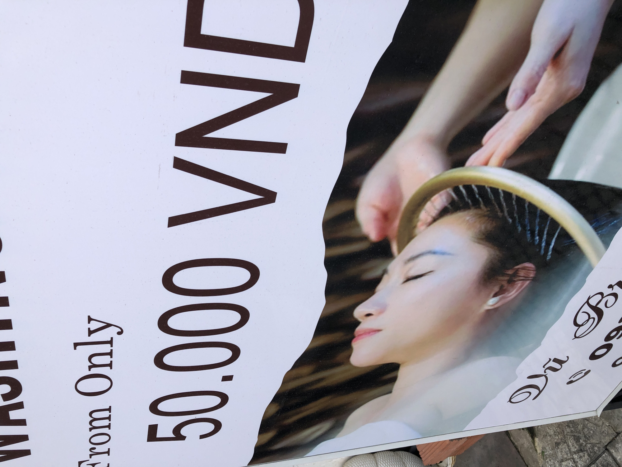 A close-up photograph shows a white sign with dark brown text and an image of a person receiving a treatment. The top of the sign reads "WASHING" in large letters. Below this, in smaller text, it says "From Only". The most prominent text on the sign is "50.000 VND", indicating a price. The lower portion of the sign features a photograph of a person lying down, with their head tilted back and eyes closed, appearing to be in a spa or salon. Their hair is being tended to by hands, suggesting a hair washing or treatment service. The person's skin is fair, and they have red lipstick on. Below the image of the person, there is text in a script font, partially obscured by shadows, with "Vũ" being the only clearly legible word. The sign is displayed outdoors, as indicated by the pavement and a wooden bench visible in the lower left and bottom of the image. The location is identified as Đà Nẵng, Vietnam.