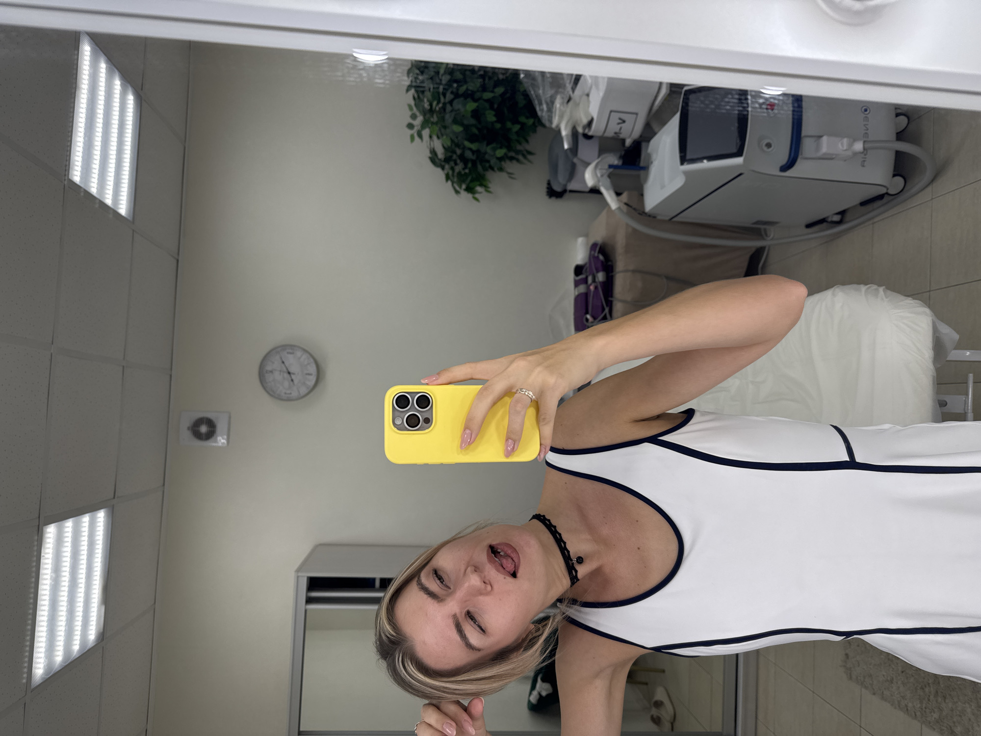 A young woman is taking a selfie in a room. The room is likely a medical or beauty treatment room, based on the medical device visible in the background, which is white and blue. The room also contains a plant, a clock on the wall, and a padded treatment table. The woman is holding a yellow smartphone, her face is close to the camera, and she is sticking out her tongue. She is wearing a white dress with navy trim. The location appears to be indoors, possibly in a professional setting.