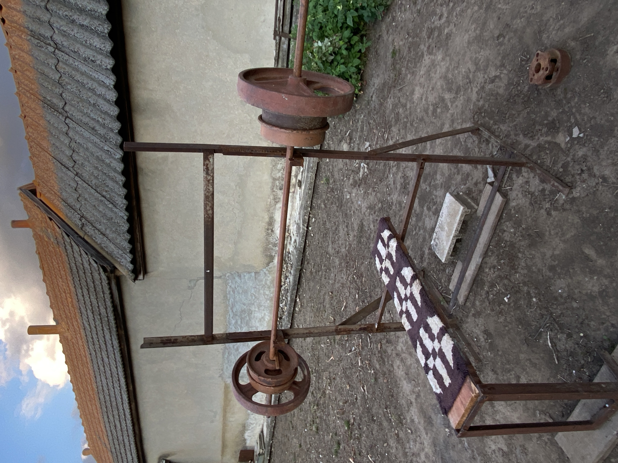 The image shows an outdoor workout bench with a barbell and weights, situated in a yard with a dirt ground. The bench is made of rusted metal and has a small, patterned mat on the seat. The barbell is loaded with two large, circular weights on each side. In the background, there is a building with a tiled roof and a stucco wall. To the right of the bench, there is green foliage. The sky above is partly cloudy, with the sun casting a warm light on the scene. There are no people visible in the image. The scene appears to be in a rural or semi-rural setting. This image was taken in Cairaclia, Moldova.