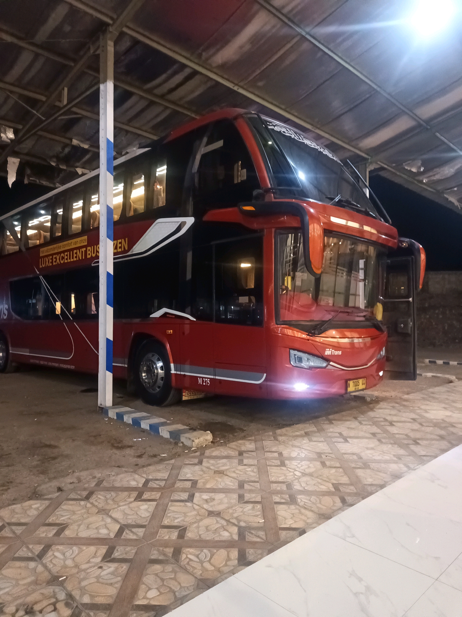 A red double-decker bus with the text "LUXE EXCELLENT BUSZEN" and "HT Trans" is parked under a covered structure. The bus is illuminated by its headlights and interior lighting, revealing its upper and lower decks. The bus is situated on a patterned tiled surface, with a portion of the ground being dirt. Several blue and white striped concrete blocks are placed in front of the bus. The environment appears to be a bus terminal or a rest stop, possibly in Cikawung, Indonesia. There are no people visible in the image.