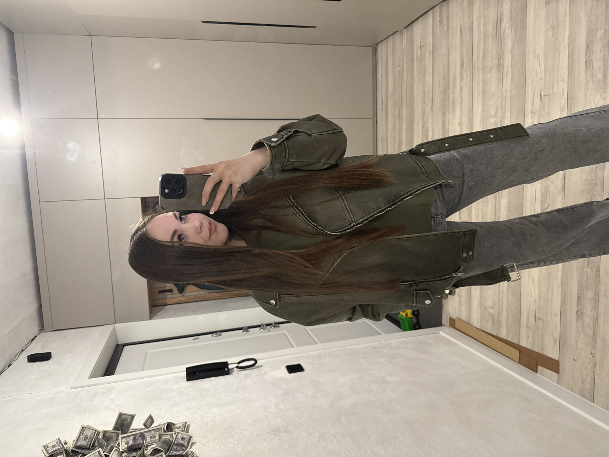 A woman stands in a room, holding a smartphone up to take a selfie in front of a mirror. She is wearing a green jacket and gray jeans. The background features a wardrobe, wood-like flooring, and a white door. There is a pile of US dollar bills on the floor. The location is likely an apartment or home. The city is not discernible from the image.