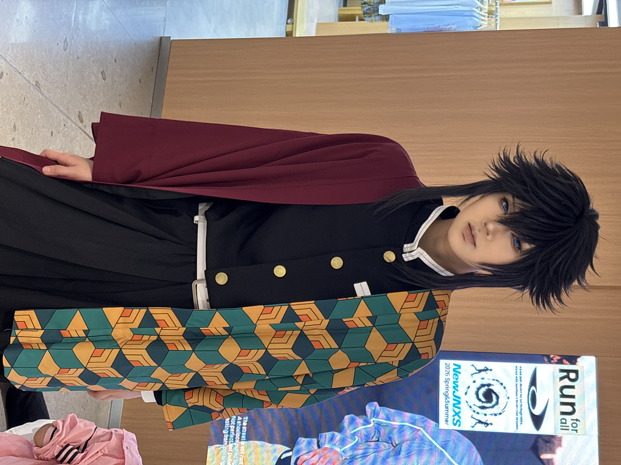 A person dressed as the character Giyu Tomioka from the anime series "Demon Slayer" stands indoors. The person is wearing a black uniform with gold buttons, a red cloak, and a green and orange patterned haori. They have black hair and blue contact lenses. The location is likely a retail environment in Liangjiang, China, although that cannot be verified with 100% certainty given the limited visual information. A promotional poster for a "Run for all" event is partially visible nearby.