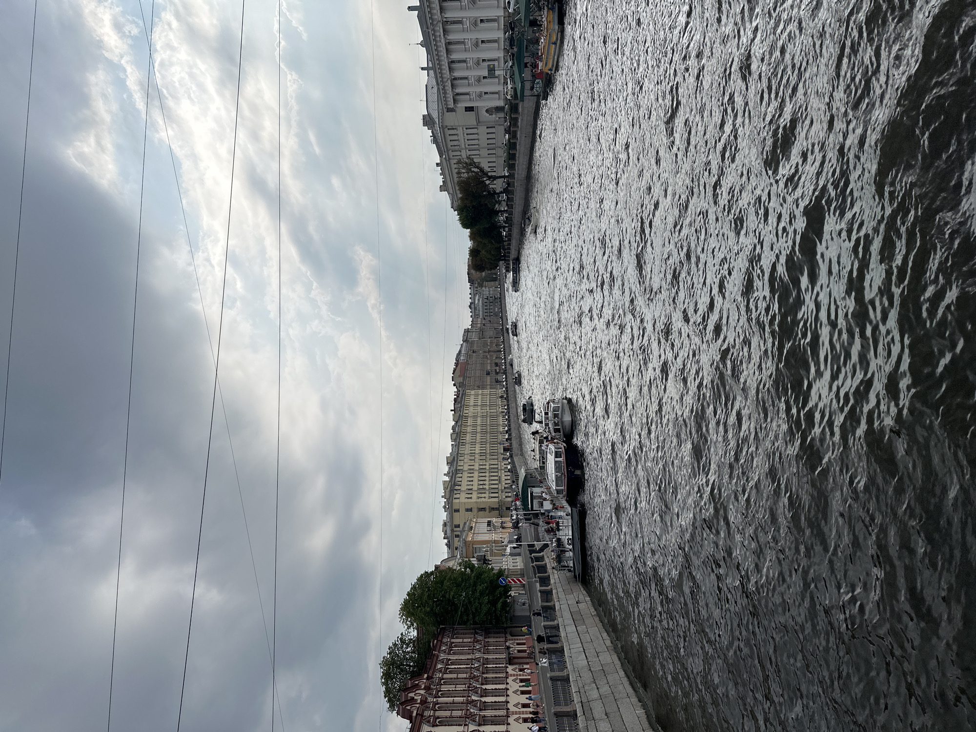 The image depicts a wide canal with a cloudy sky overhead. The canal is lined with historic buildings on both sides, characteristic of Saint Petersburg, Russia. Several boats, including passenger ferries and smaller vessels, are visible on the water. People are seen walking along the canal's embankment and boarding the boats. The architecture suggests a European city with a rich history, likely a prominent waterway within Saint Petersburg, possibly the Moyka River or the Fontanka River, given the style of the buildings and the presence of tour boats. The water's surface is rippled, indicating a gentle current or breeze. The overall atmosphere is overcast, suggesting a typical Northern European summer day.