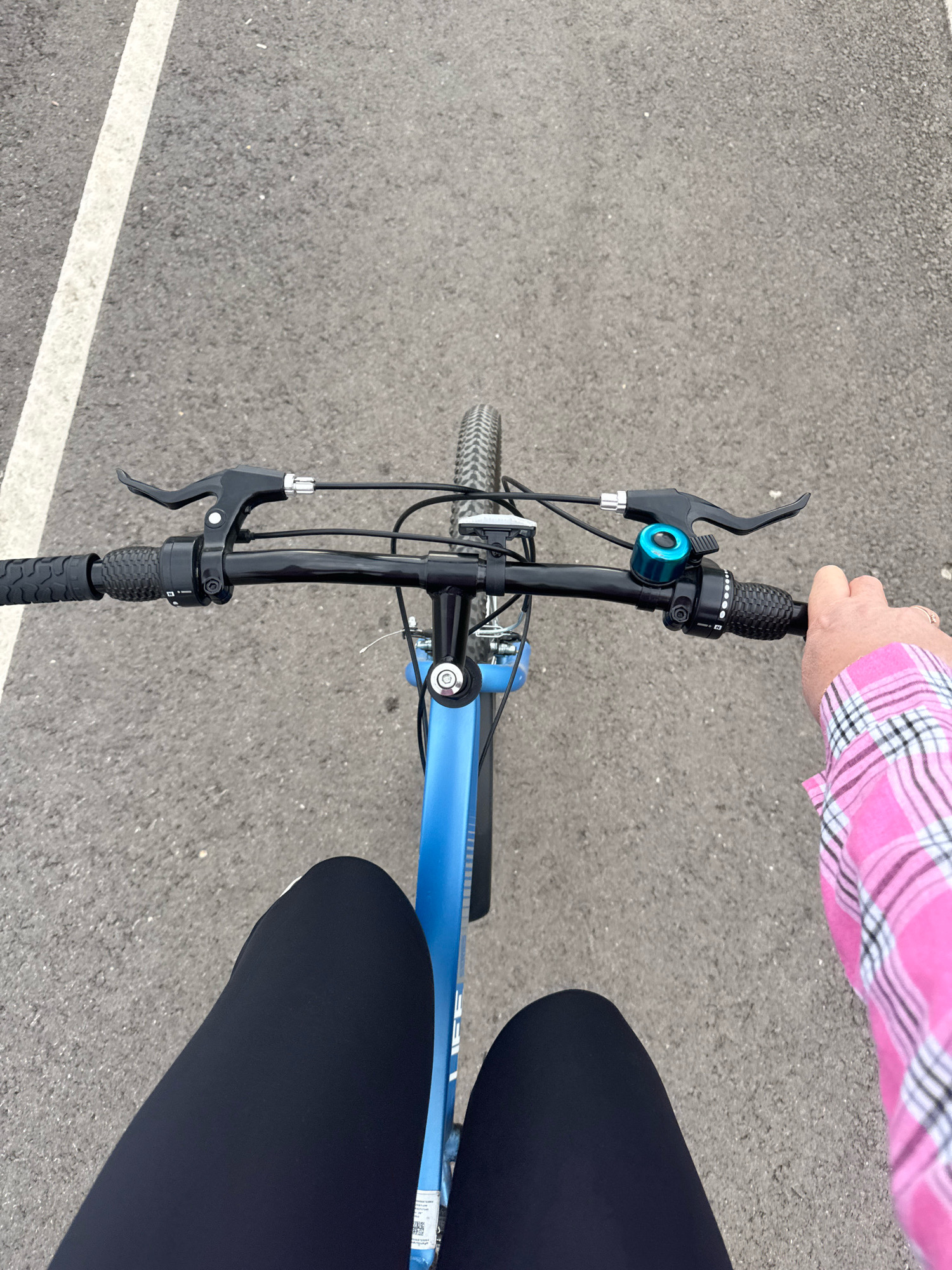 This is a first-person point-of-view shot of a person riding a bicycle on an asphalt road. The road has a white painted line on the left side. The bicycle is blue. The rider is wearing black leggings and a pink and white plaid shirt. The rider's left hand is on the handlebar, and the right hand is holding the right handlebar grip. A blue bicycle bell is attached to the handlebars. The image does not provide any visual clues to determine the specific location in Ufa, Russia.