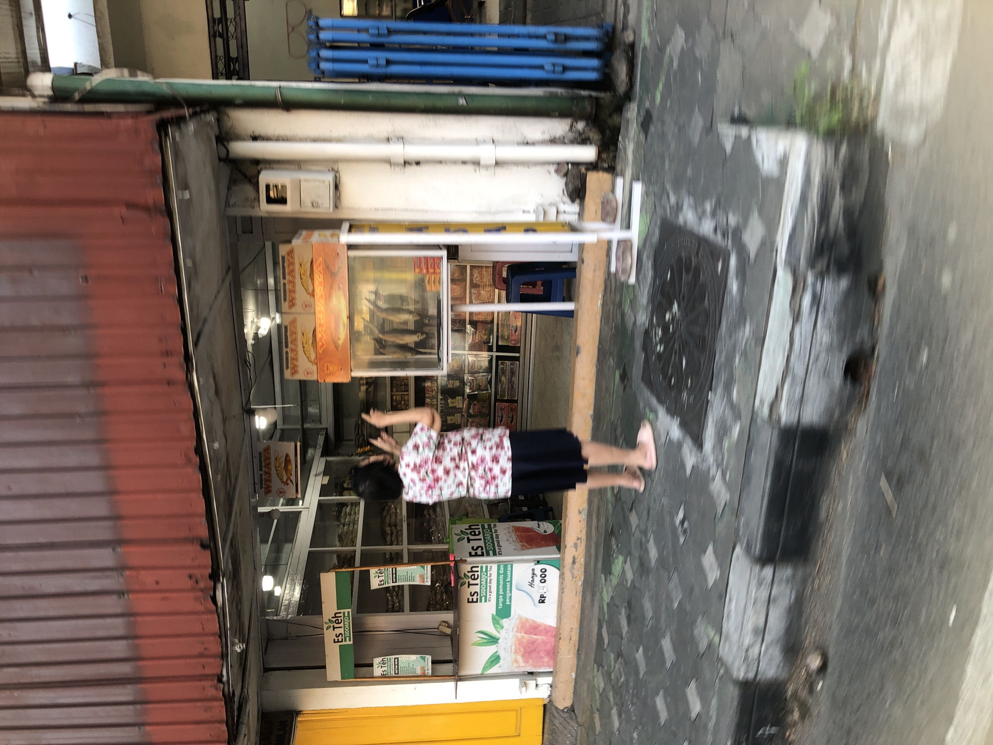 A person, appearing to be a woman with dark hair, stands facing away from the camera, gesturing with both hands towards a storefront. She is wearing a floral print shirt and a dark skirt, with light-colored sandals on her feet. The storefront is open, revealing shelves stocked with various products, some of which appear to be packaged goods. In front of the storefront, there is a display case with hanging items, possibly food. To the left of the storefront, a yellow structure is visible, and to the right, blue folding doors are present. The pavement in front of the store is paved with square tiles. The scene appears to be a street in Sidoarjo, Indonesia, based on signage that reads "Es Teh Sidoarjo."