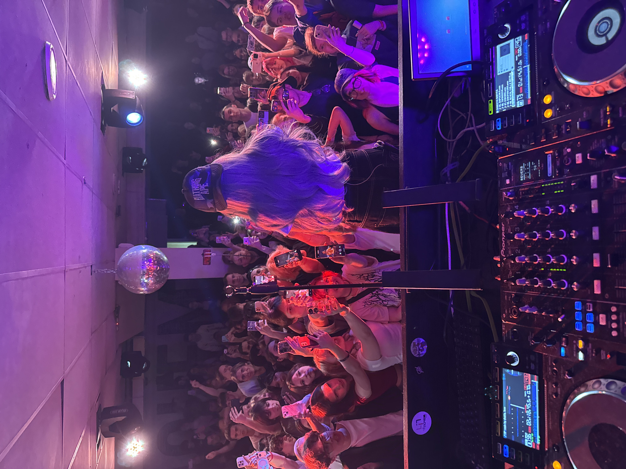 The image shows a crowd of people at a live music event, with a DJ performing on stage. Many audience members are holding up their phones, presumably recording or taking pictures. A disco ball hangs from the ceiling. The DJ equipment is prominently displayed. The stage lighting is vibrant, featuring blue and purple hues. The location is Krasnoyarsk, Russia, as inferred from the Cyrillic text "ЧЕПОЧЕМ" and "КРАСНОЯРСК" sticker visible on DJ equipment. The room appears dimly lit, other than the stage lights and camera flashes.