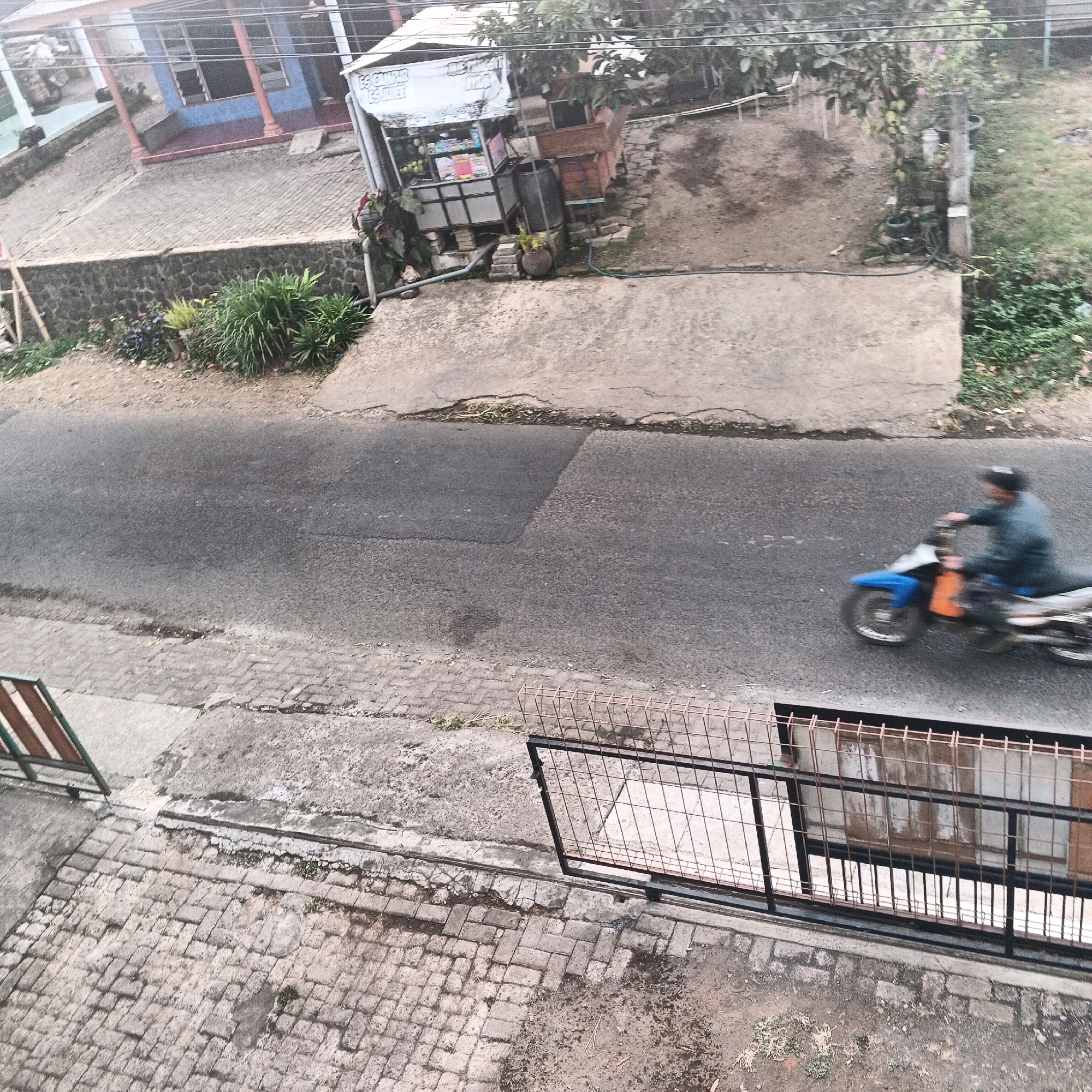 This high-angle shot captures a street scene in Bocek, Indonesia, likely during daylight hours. The asphalt road dominates the mid-ground, with a single motorcyclist in motion on the right side. The motorcyclist, wearing a blue jacket and a helmet, is blurred due to motion, riding an orange and blue motorcycle. To the left of the road, a paved sidewalk transitions to a stone retaining wall, beyond which a house with a blue facade and red pillars is visible. In the background, a small food stall with a white awning is situated, displaying various items for sale. Further back, more houses and vegetation are present. The foreground shows a brick-paved area with a metal fence. The environment appears somewhat weathered, with visible cracks in the pavement and a generally dry ground surface, suggesting a possibly warm climate.