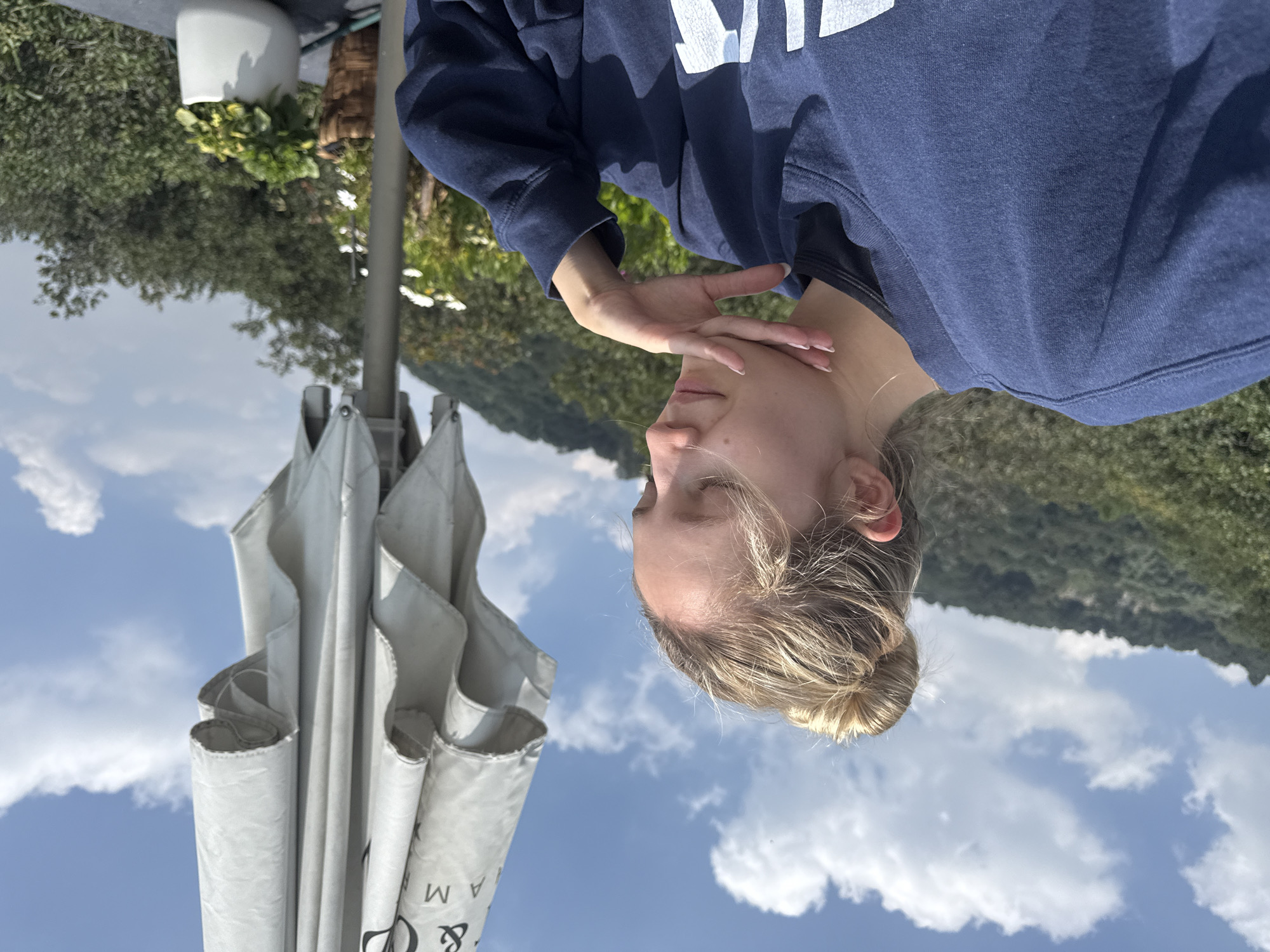 A young woman with blonde hair in a bun sits outdoors with her eyes closed and her hand on her chin. She is wearing a navy blue sweatshirt. Behind her, a large, closed umbrella is visible, with partial text that reads " & AM". The background shows a lush, green mountainside under a blue sky with white clouds. The setting appears to be a restaurant or cafe terrace with potted plants visible in the foreground. The image suggests a peaceful moment of reflection or relaxation in a scenic location. The presence of the umbrella and the style of the restaurant suggests it could be in the Polanco neighborhood of Mexico City, known for its upscale dining and cafes.