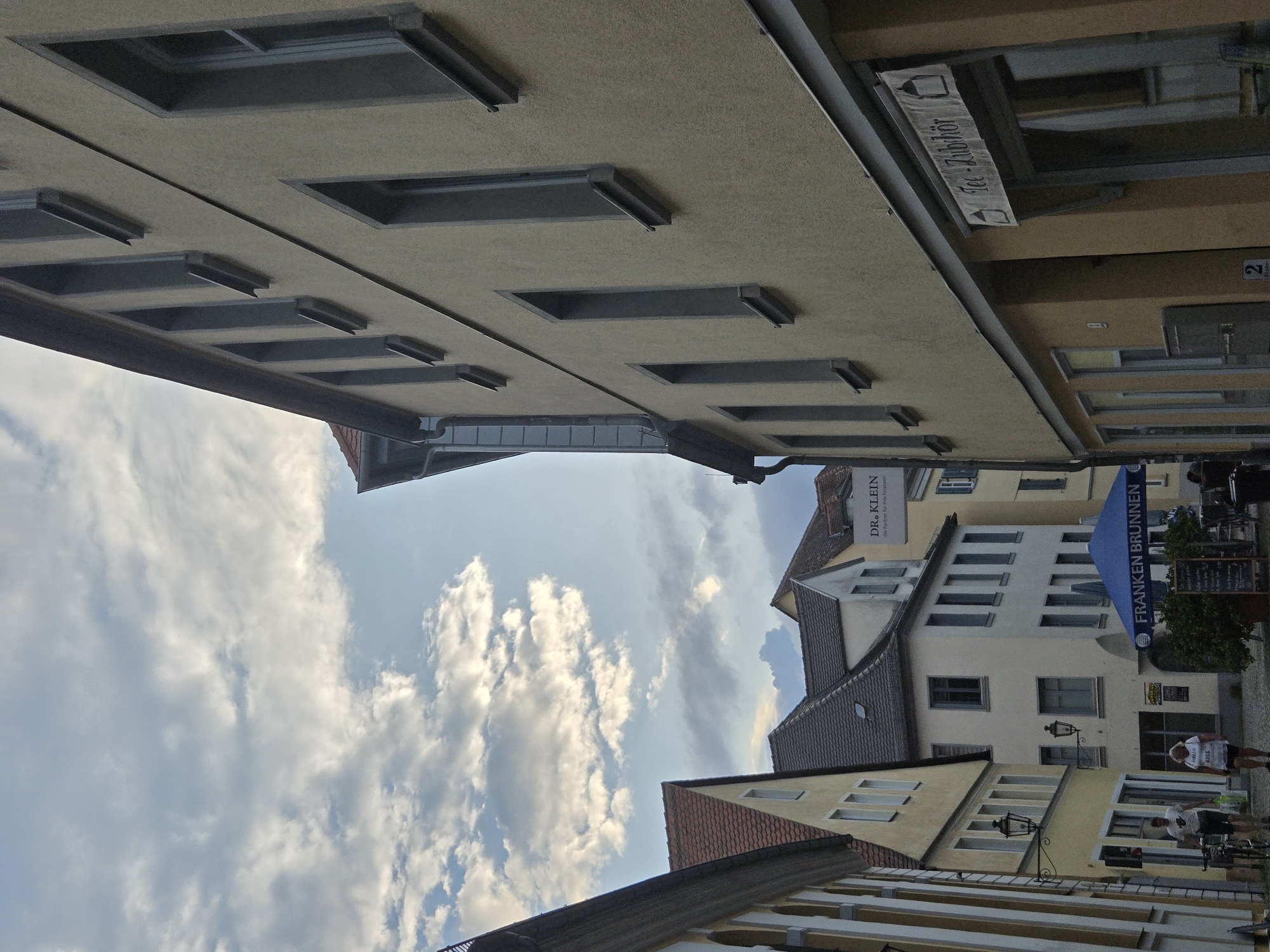 This is a photograph of a street scene in Ansbach, Germany, taken from a low angle looking upwards. The buildings are old and have traditional architecture, with tiled roofs and cream-colored facades. The sky is filled with fluffy white clouds.
In the foreground, there are several modern awnings extending from the buildings. One of these awnings is blue and has text that reads "FRANKEN BRUNNEN". Another awning has the text "DR. KLEIN" and "Die Partner für thee Finanzen". There is also a banner that appears to say "Tec-Zubhör" with a graphic of a tea cup.
In the background, there are people walking on the street, suggesting it is daytime and likely a busy period. One person is wearing a white t-shirt with the word "HELLO" and "KISS" on it. There are bicycles parked outside one of the buildings. The lighting suggests it is a bright, possibly sunny day, but the presence of many clouds indicates overcast conditions or a partly cloudy sky. The overall impression is a charming European town with a mix of old and new elements.