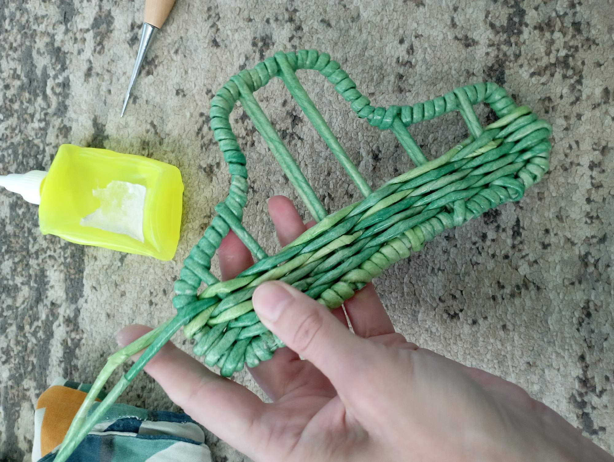 A close-up, overhead shot shows a person's hand holding a green, woven object. The object appears to be a basket or a decorative piece, made from rolled and twisted paper or natural fibers. The weaving is intricate, with individual strands of varying shades of green creating a textured surface.
The hand is positioned over the woven object, with fingers gently supporting it from above. The skin tone of the hand is light, and the fingernails are short and clean.
The object is placed on a textured surface that resembles a carpet or a textured fabric. The background is a muted beige with darker speckles, giving the impression of a worn or rustic material.
To the right of the woven object, there is a bright yellow squeeze bottle of glue. The bottle has a white cap and a clear nozzle. A small amount of white glue is visible on the label of the bottle.
Below the glue bottle and to the right of the woven object, a pointed metal tool with a wooden handle is visible. The tool appears to be an awl or a similar crafting implement, used for poking or guiding materials.
In the upper right corner, a portion of a folded piece of fabric with a geometric pattern in shades of blue, green, and orange is visible, partially obscuring the background.
The scene is illuminated by overhead lighting, casting soft shadows. There is no discernible information within the image that would associate it with Blagoveshchensk, Russia.
