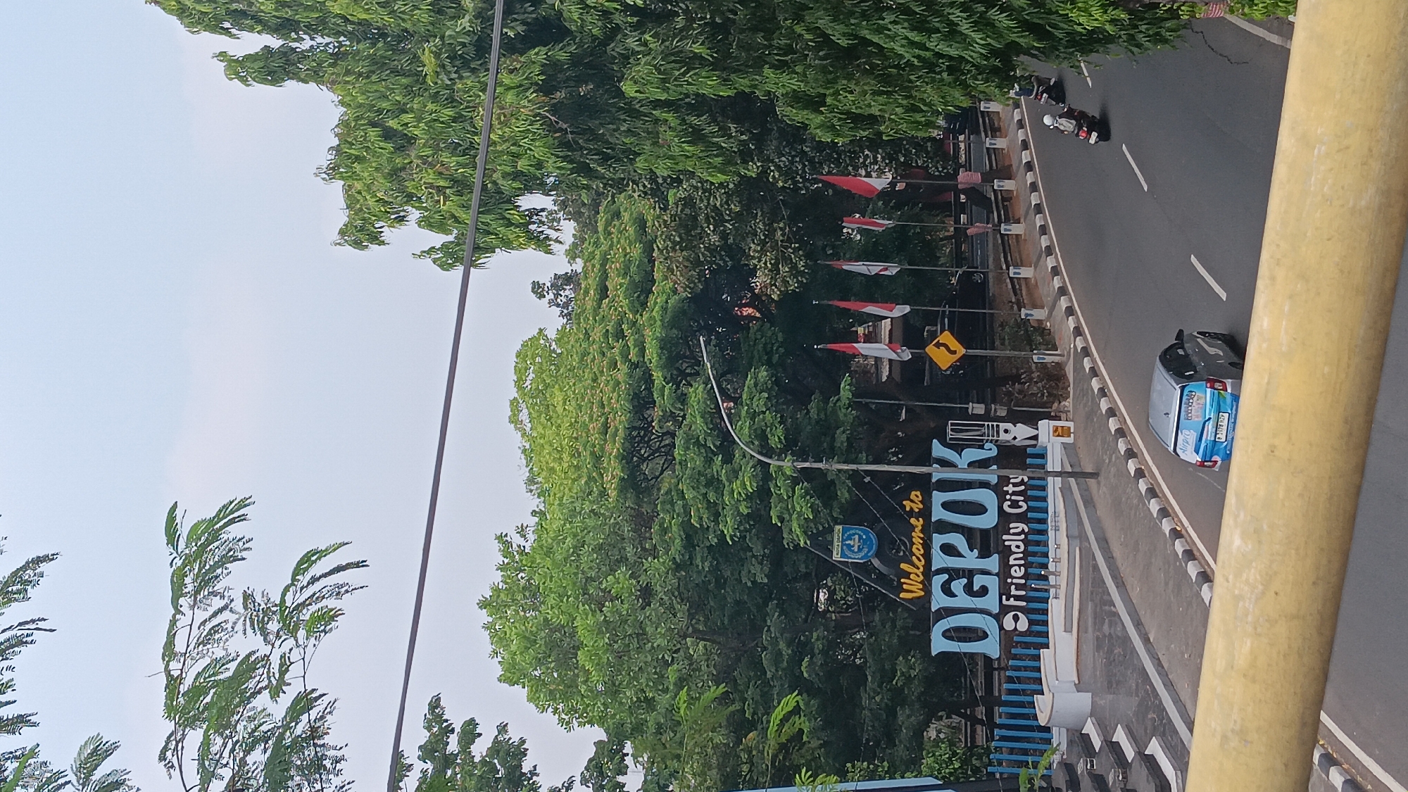 The image shows an outdoor scene in Depok, Indonesia. A large, blue sign with "Welcome to DEPOK Friendly City" is prominently displayed, featuring the city's emblem. Several Indonesian flags are planted behind the sign. The road in front of the sign curves to the right, with a silver car and two motorcycles driving on it. Lush green trees surround the area, with a cloudy sky overhead.