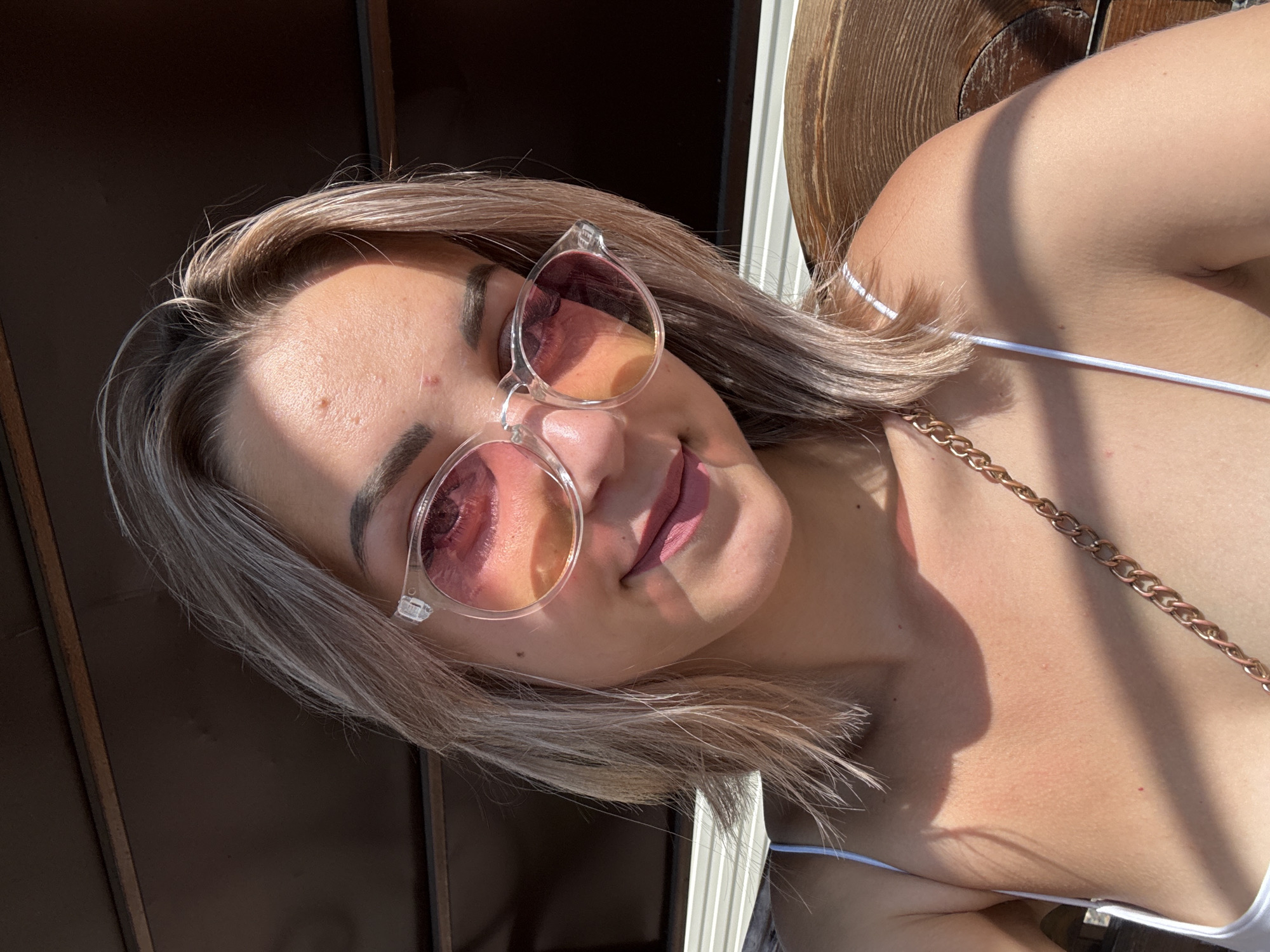 A woman with blonde shoulder-length hair and pink-tinted sunglasses smiles at the camera. She is wearing a white camisole and a gold chain purse strap is visible. She has makeup on, including lipstick. The background appears to be outdoors with wooden structures and dappled sunlight. The image is a selfie, taken from the chest up. The setting is not identifiable as Krasnogorskoye, Russia.