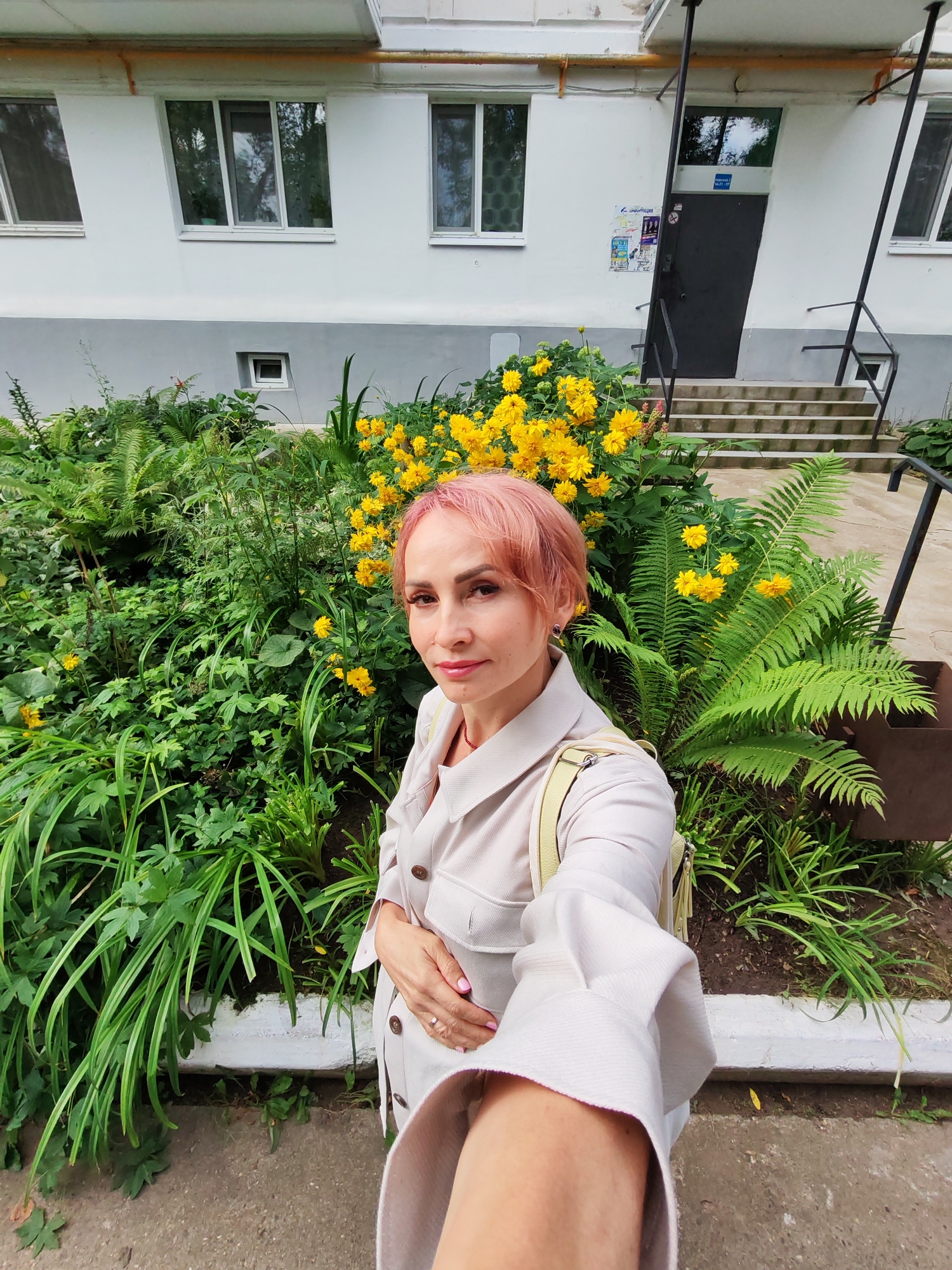 A woman with short pink hair and a light-colored trench coat takes a selfie outdoors in front of a building with yellow flowers and lush green foliage. She is wearing a ring on her left hand and has a light yellow bag slung over her shoulder. Behind her, a white building has several windows and a dark entrance door with a set of stairs leading up to it. A sign with Cyrillic text is affixed to the wall next to the door. The location is in the city of Chaikovsky, Russia, as indicated by the presence of a sign with Cyrillic text and typical Soviet-era apartment building architecture.