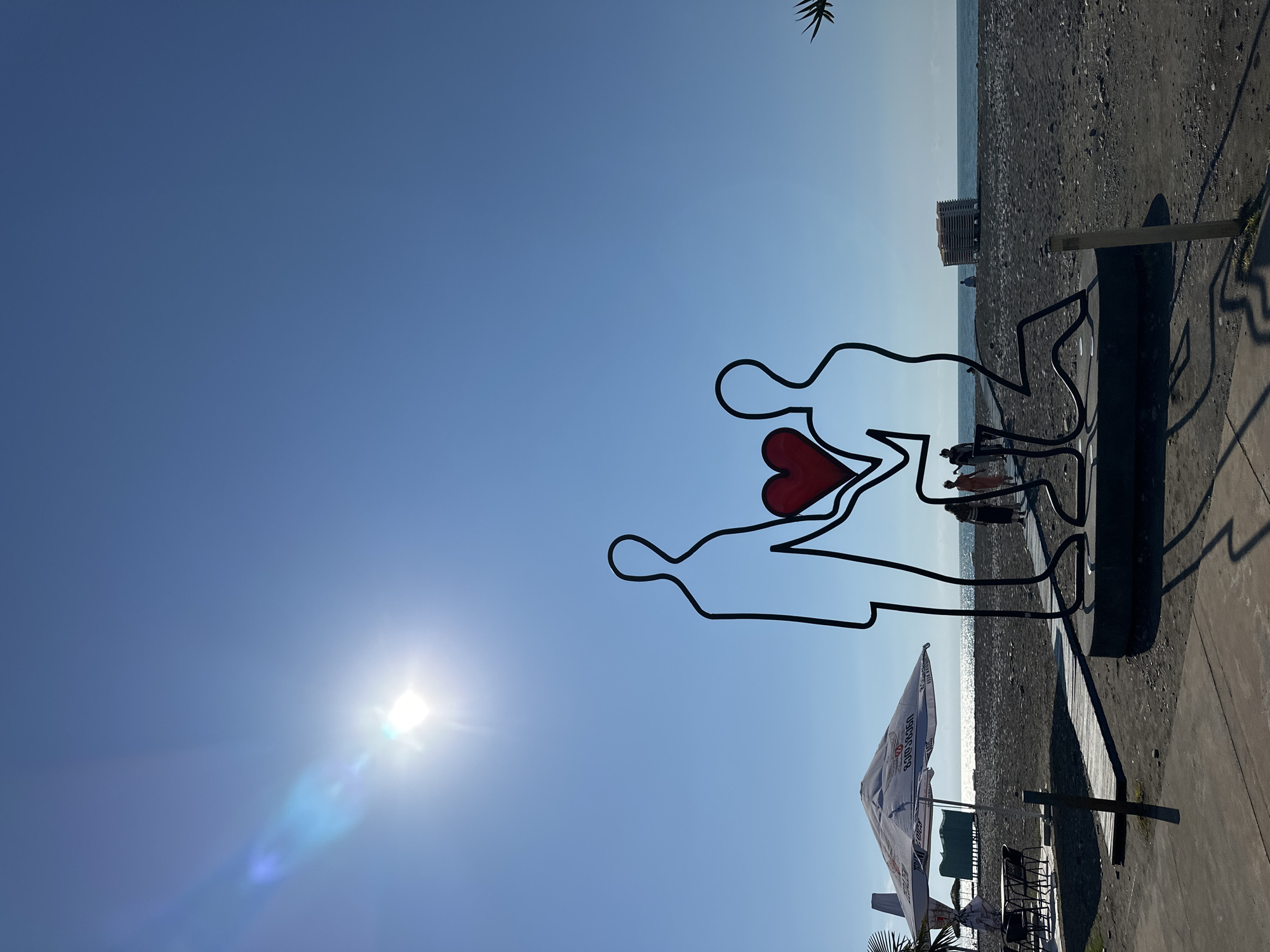 An outdoor sculpture of two abstract figures, one kneeling and holding a red heart towards the other standing figure, is positioned on a black pebble beach in Batumi, Georgia. The sun shines brightly in a clear blue sky, casting shadows from the sculpture and a nearby umbrella. In the background, the calm sea stretches to the horizon, with a few people walking along the beach and a stack of lounge chairs. To the left, a white umbrella shades tables and chairs of an outdoor cafe. The sculpture appears to be a romantic gesture, possibly a proposal.