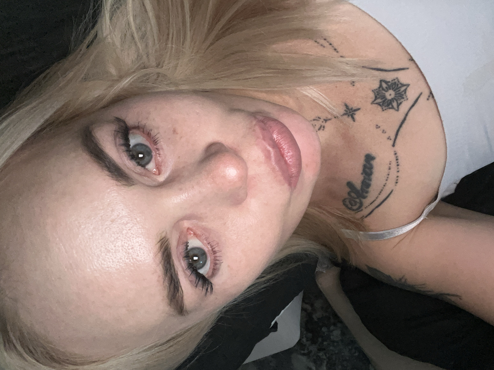 This is a close-up, slightly angled, overhead shot of a young, fair-skinned woman with blonde hair and blue eyes. She is lying down, looking upwards towards the camera. Her lips are slightly parted and appear to be wearing lipstick. She has on false eyelashes and what appears to be light makeup.
She has several tattoos visible on her shoulder and collarbone area. One tattoo on her shoulder is a geometric, eight-pointed star shape. Above and to the left of this are two smaller star shapes. Below the star tattoo, on her collarbone, is a word in script that appears to read "Aurora". Further down her chest, a thin strap of what looks like lingerie is visible. Another tattoo is visible on her upper arm, appearing to be a feather or wing design.
The background is dark and out of focus, suggesting an indoor setting with low lighting. The overall mood is intimate and casual. There is no discernible time of day or weather information available from the image.