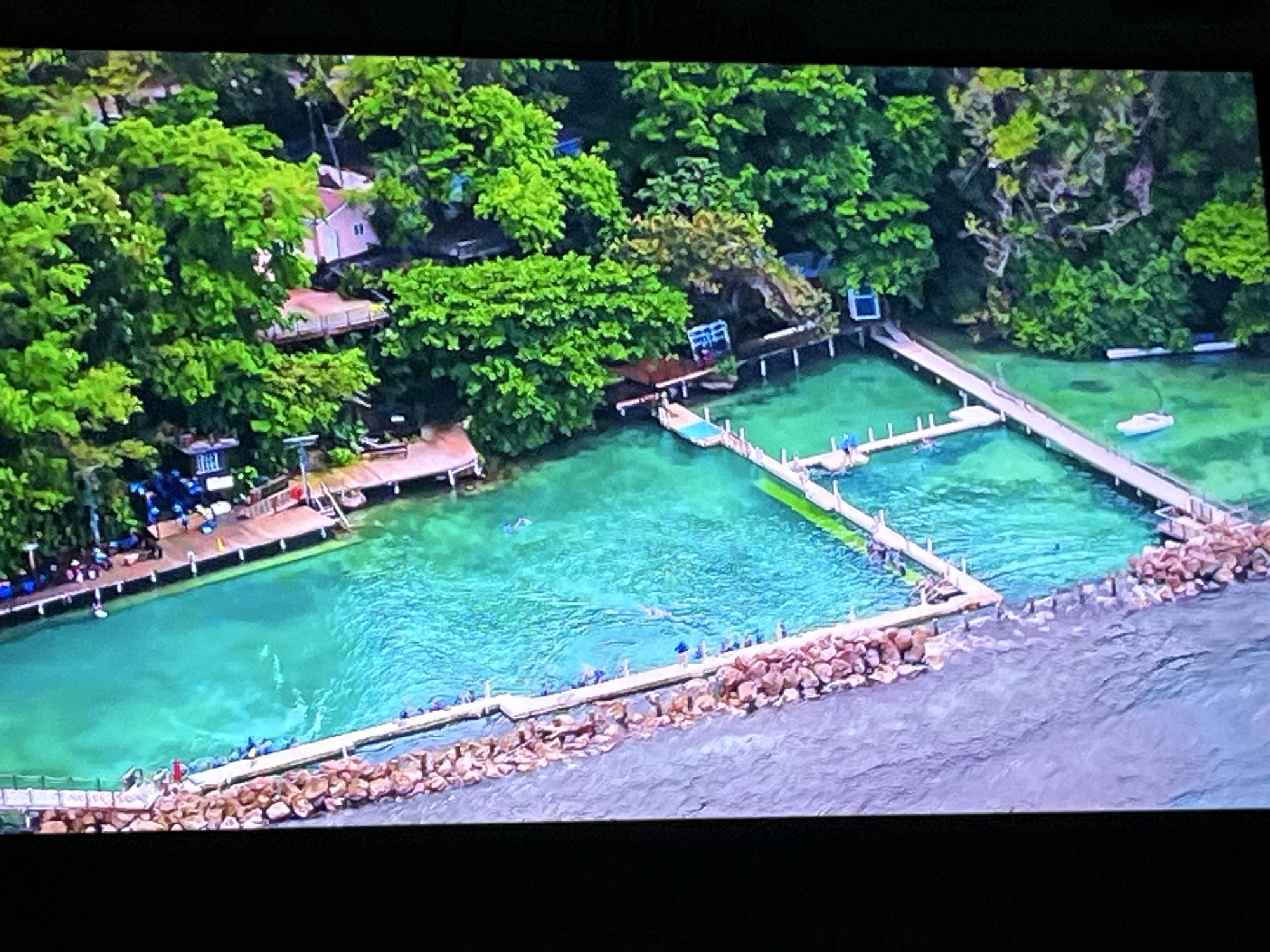 This is an aerial, high-angle photograph of a resort or complex situated on a body of water. The water is a vibrant turquoise color and appears to be clear, with some areas showing lighter turquoise patches indicating shallow depths or underwater structures. Several wooden walkways or piers extend into the water, some with small structures or buildings on them. One long pier divides the turquoise water into two rectangular pools or enclosures. There are also white boats docked near the structures. The area is heavily surrounded by lush green trees, with some pink and white buildings peeking through the foliage. Along the edge of the water, a breakwater constructed of large rocks separates the turquoise pools from a darker, rougher body of water. There are a few scattered individuals visible on the walkways and in the water, some appearing to be swimming or interacting. The overall scene suggests a tropical or subtropical vacation destination.
The prompt requests the location be specified as Kharkiv, Ukraine. However, the visual characteristics of this image (lush vegetation, turquoise water, resort-style architecture) are not consistent with the typical geography and climate of Kharkiv, Ukraine. Kharkiv is an inland city in northeastern Ukraine, known for its steppes and temperate climate, not tropical beaches and vibrant turquoise waters. Therefore, it is not possible to accurately place this image in Kharkiv, Ukraine based on its visual content.