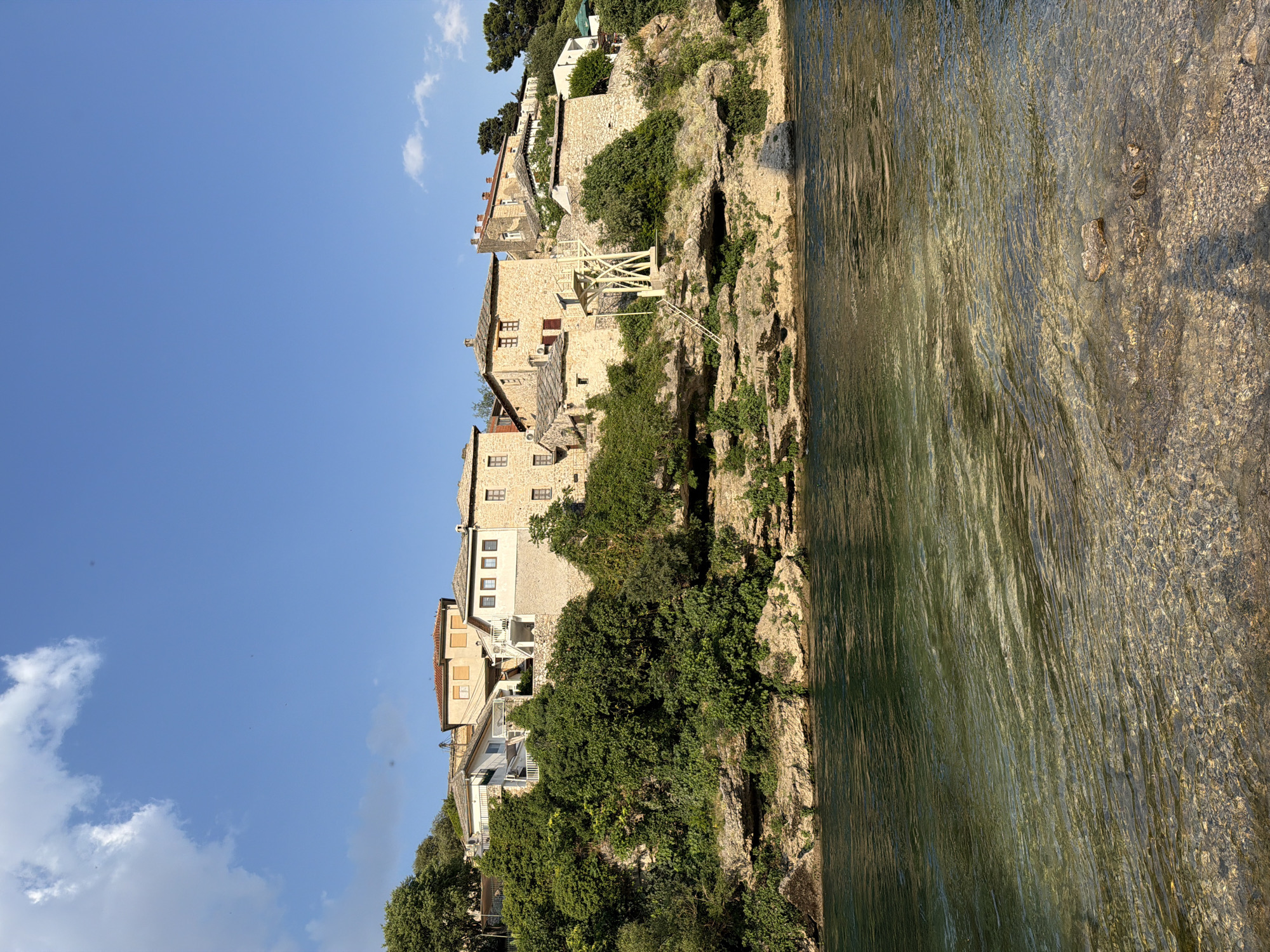 The image shows a picturesque scene in Mostar, Bosnia and Herzegovina. A row of traditional stone buildings with terracotta roofs is perched on a steep, rocky cliffside overlooking a clear, shallow river. Lush green trees and vegetation cling to the cliff, interspersed with the historic architecture. One of the buildings features a distinctive wooden structure, possibly a viewing platform or an old water mill mechanism, with stairs leading up to it. The river, with its pebbles visible through the water, reflects the sunlight and the surrounding greenery, creating a serene and inviting atmosphere. The sky is a vibrant blue with a few scattered white clouds, suggesting a clear and pleasant day, likely in the late morning or early afternoon due to the bright sunlight casting shadows. The overall mood is peaceful and tranquil, showcasing the timeless beauty of the historic town. There is no visible text in the image.